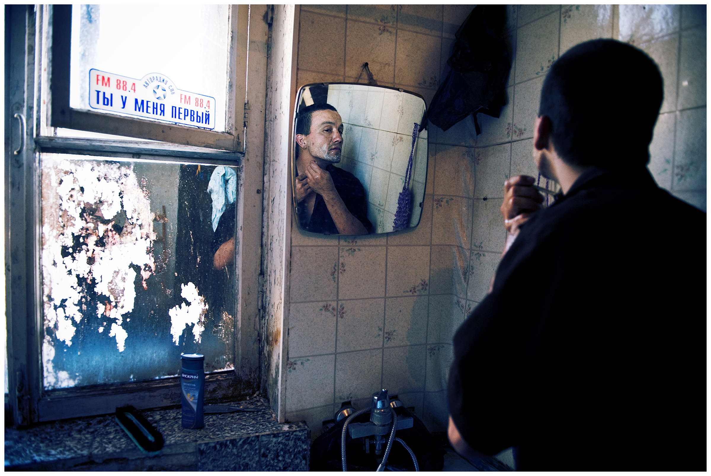 Saint Petersburg, Russia. An Uzbek guest worker shaves in the bathroom of his rented apartment. The sticker on the window has a lettering at me you are the first.