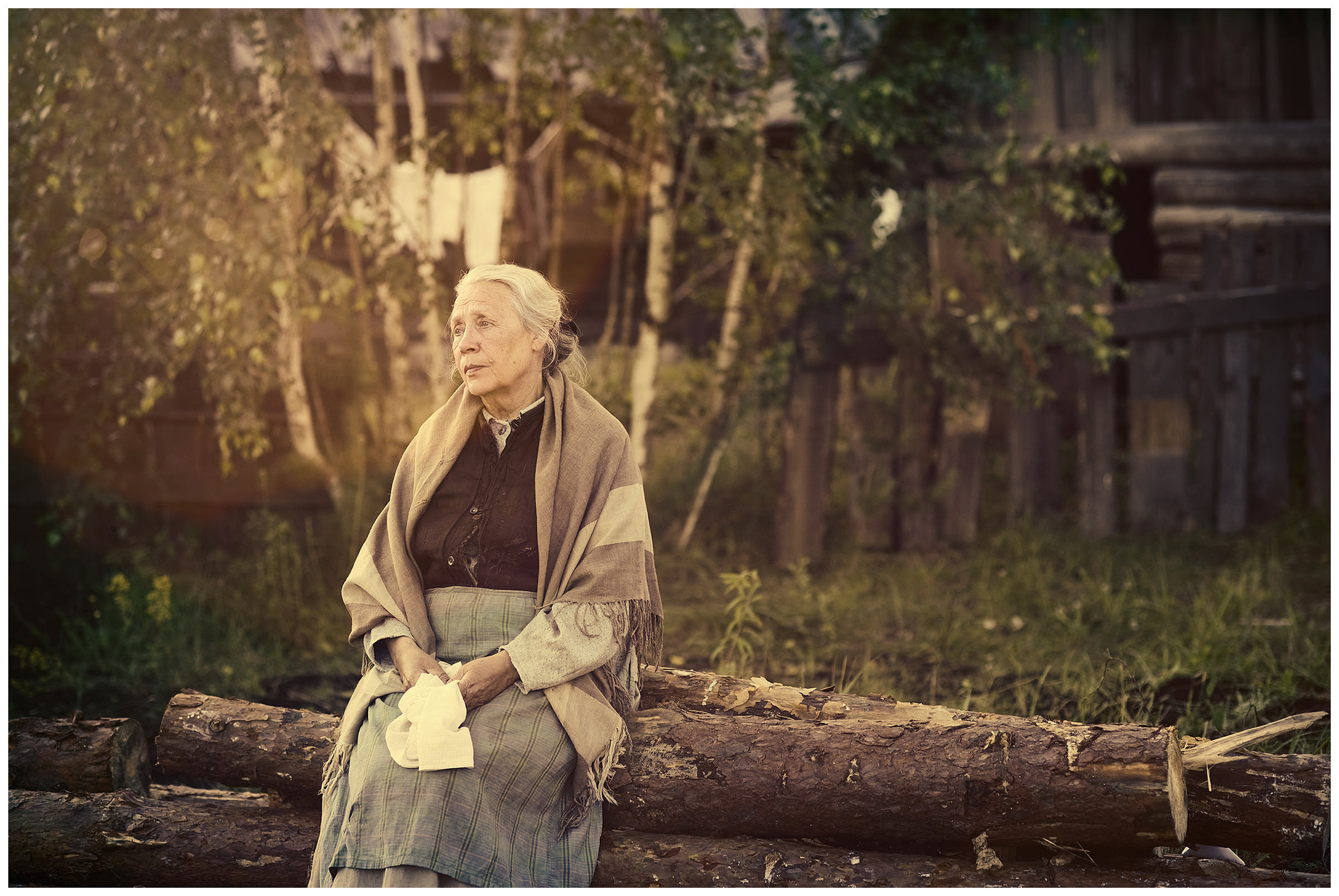 Belarus. An old woman is sitting on the tree trunks in front of a village house.