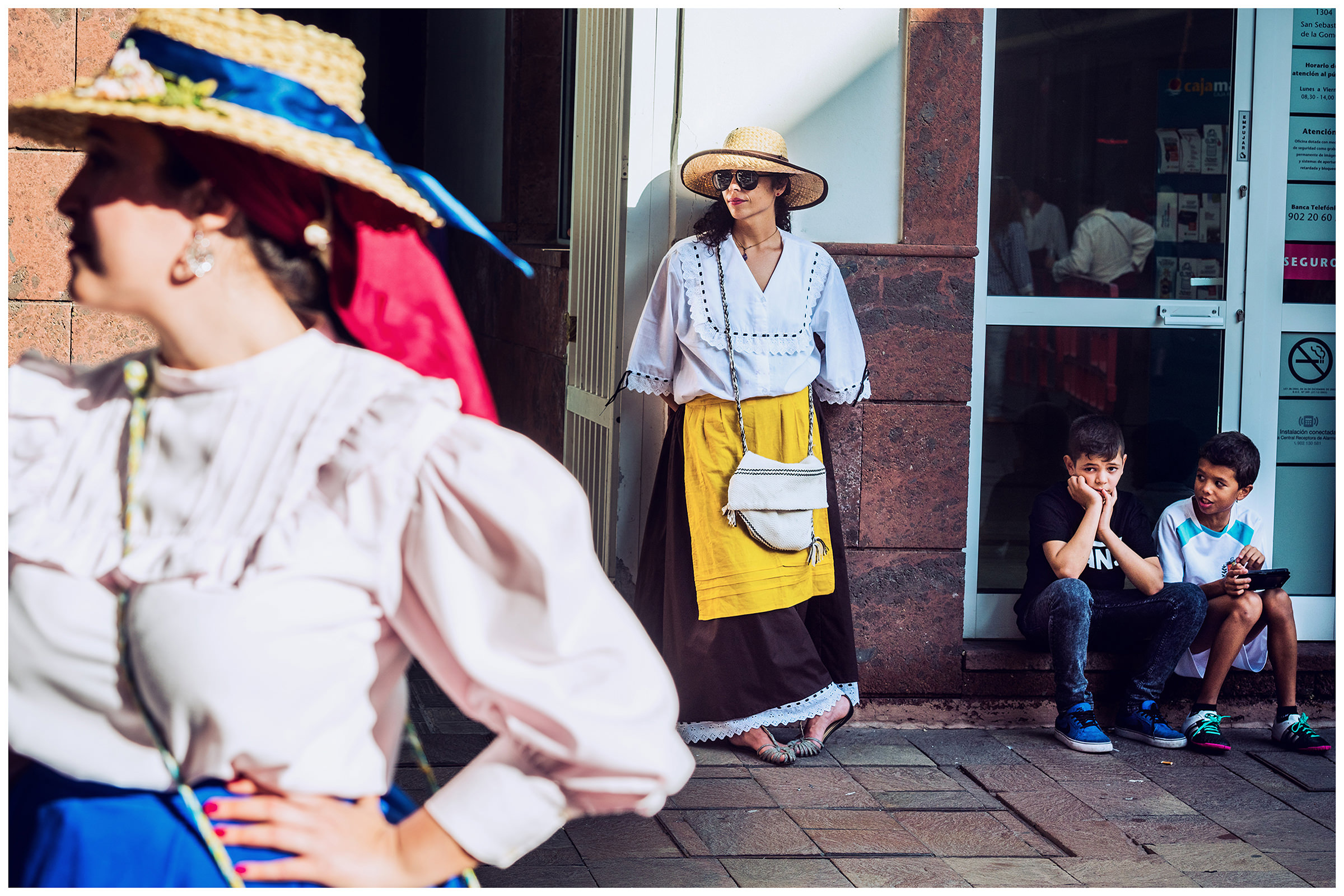 San Sebastian de la Gomera, La Gomera, Canary Islands, Spain. The inhabitants of the city of San Sebastian de la Gomera celebrate the city festival Romeria. A woman with a hat stands on a wall and two boys sit on the threshold of a shop.