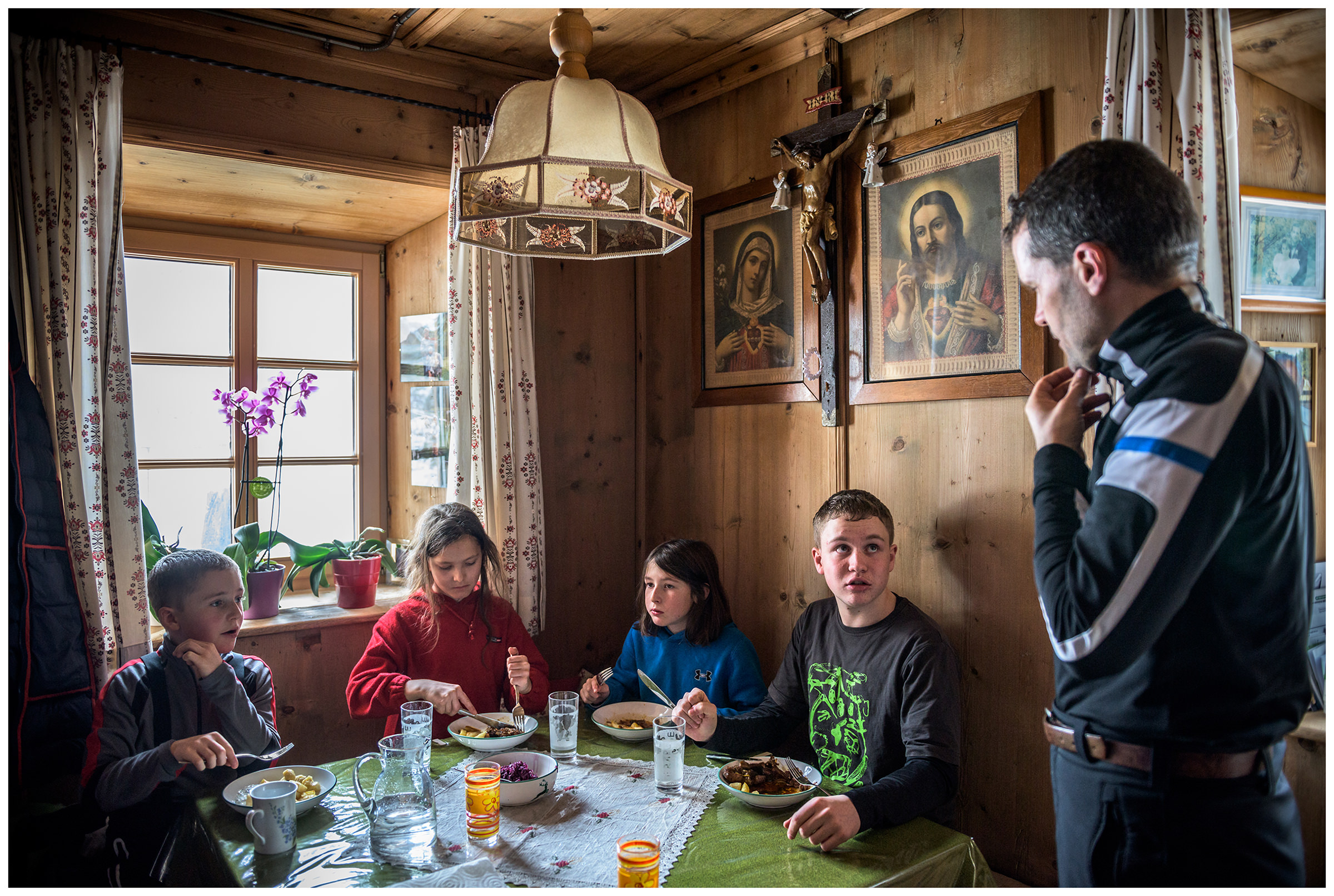 Valsertal, Wipptal, North Tyrol, Austria. Children eat at a table while a farmer talks to his son.