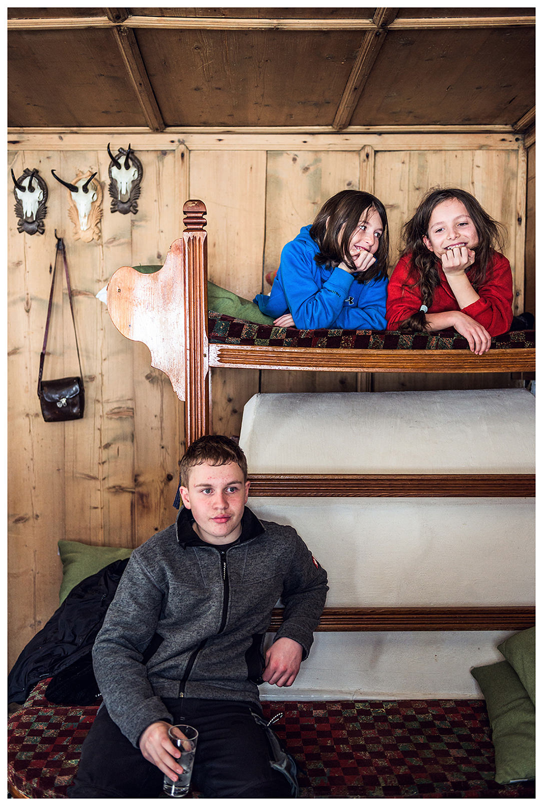 Valsertal, Wipptal, North Tyrol, Austria. Two girls lie in a bed above an oven while a boy holds a glass of water in his hand and recovers from hay drawing.