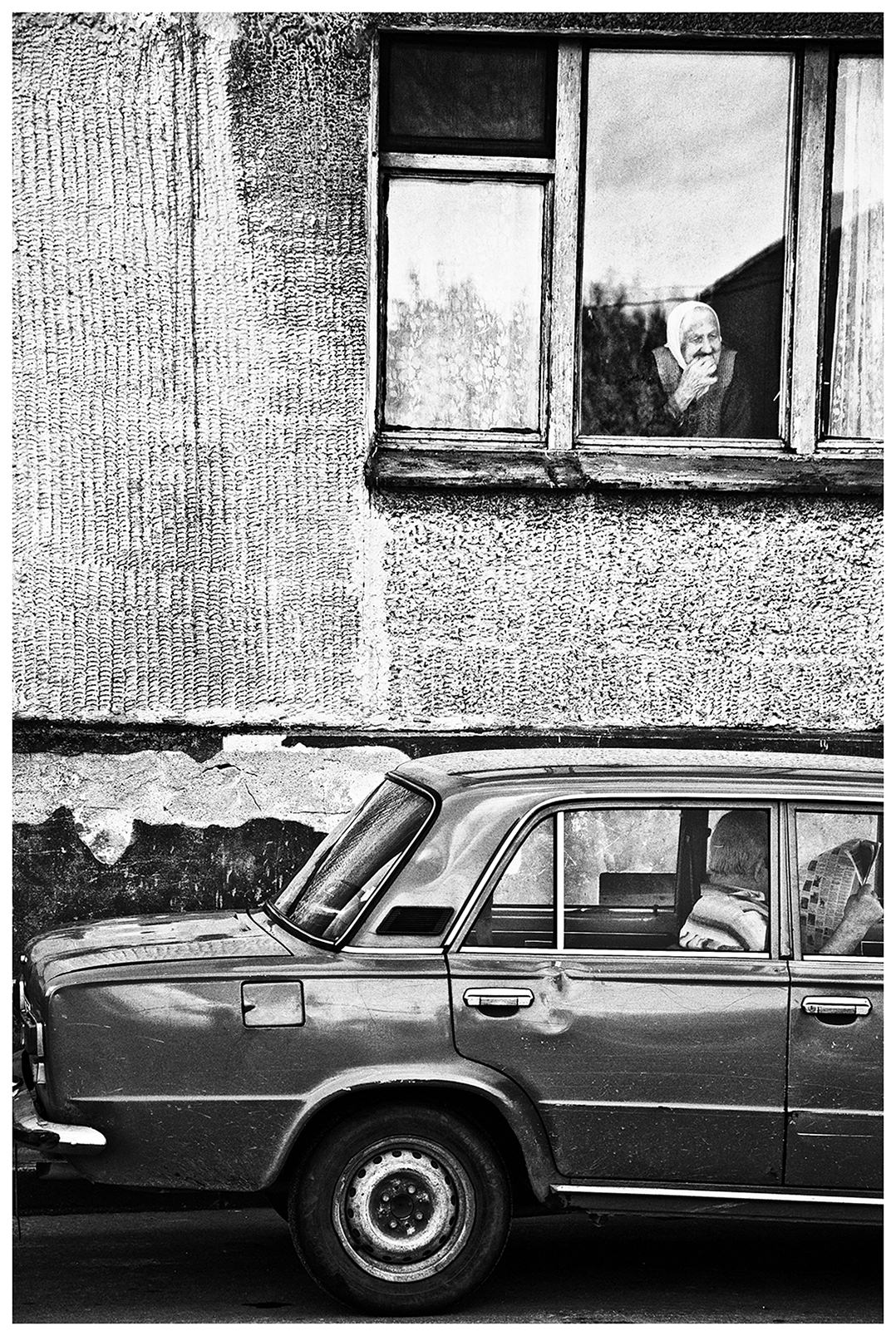 Kaliningrad, Russia. An old woman in a headscarf looks out the window while a man is sitting in a car reading a newspaper.
