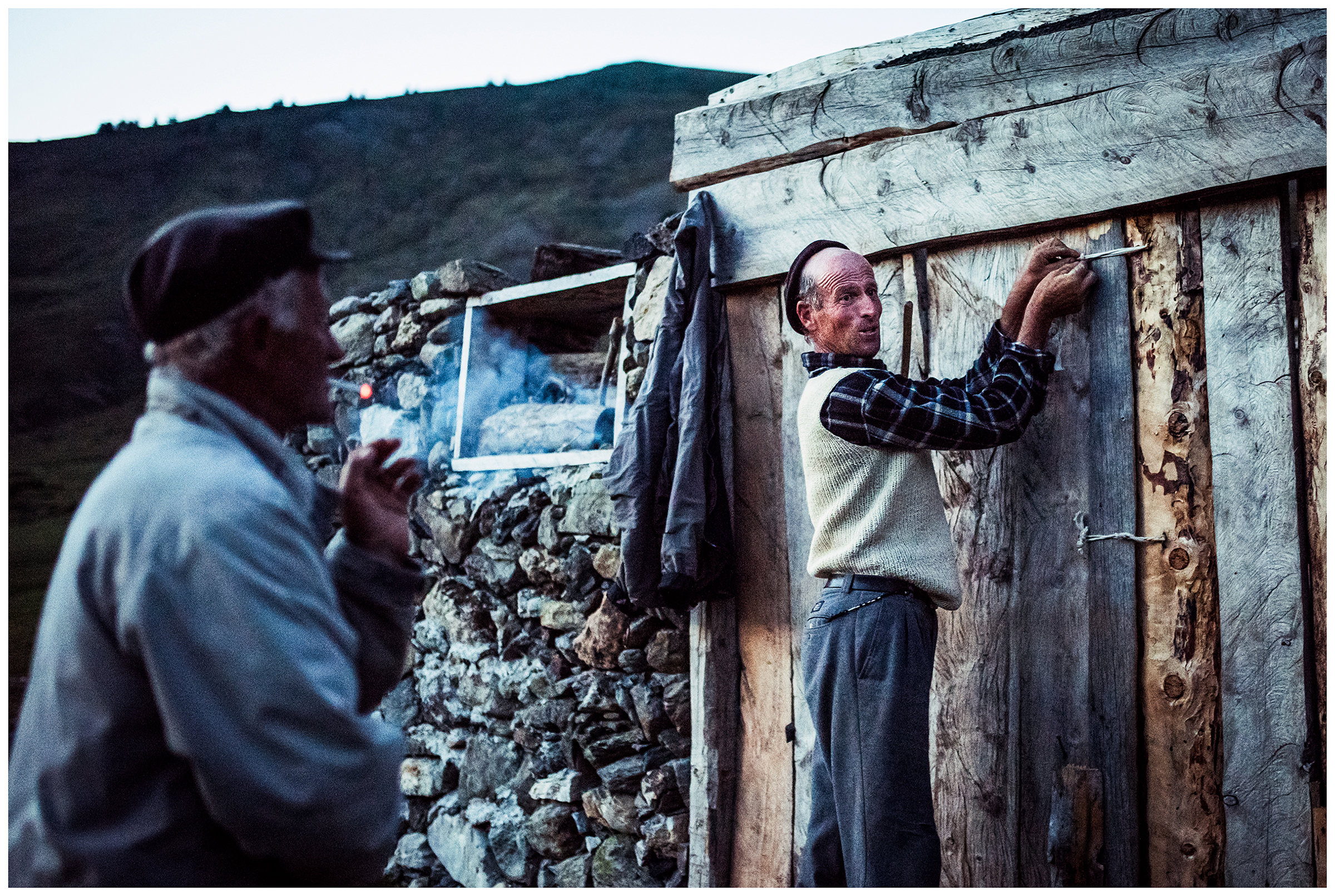 Doberdol, Albania. A villager closes a stable for the night late in the evening, while his neighbour smokes and watches him.