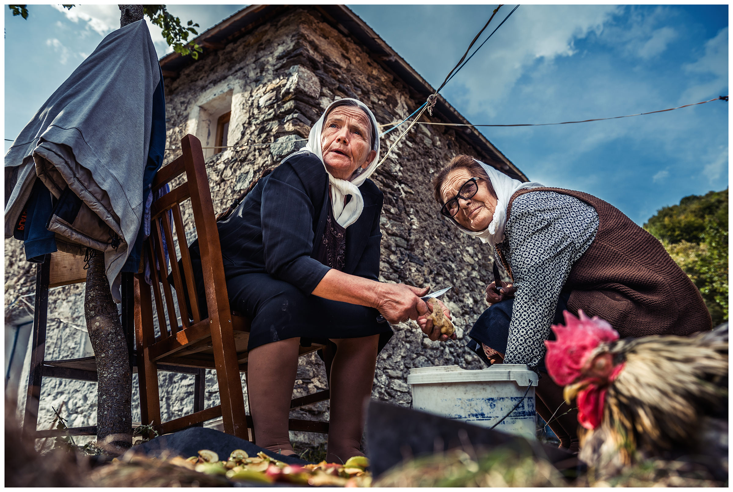 Cerem, Albania. Two old women ring potatoes in a mountain village. A cock is sitting in front.