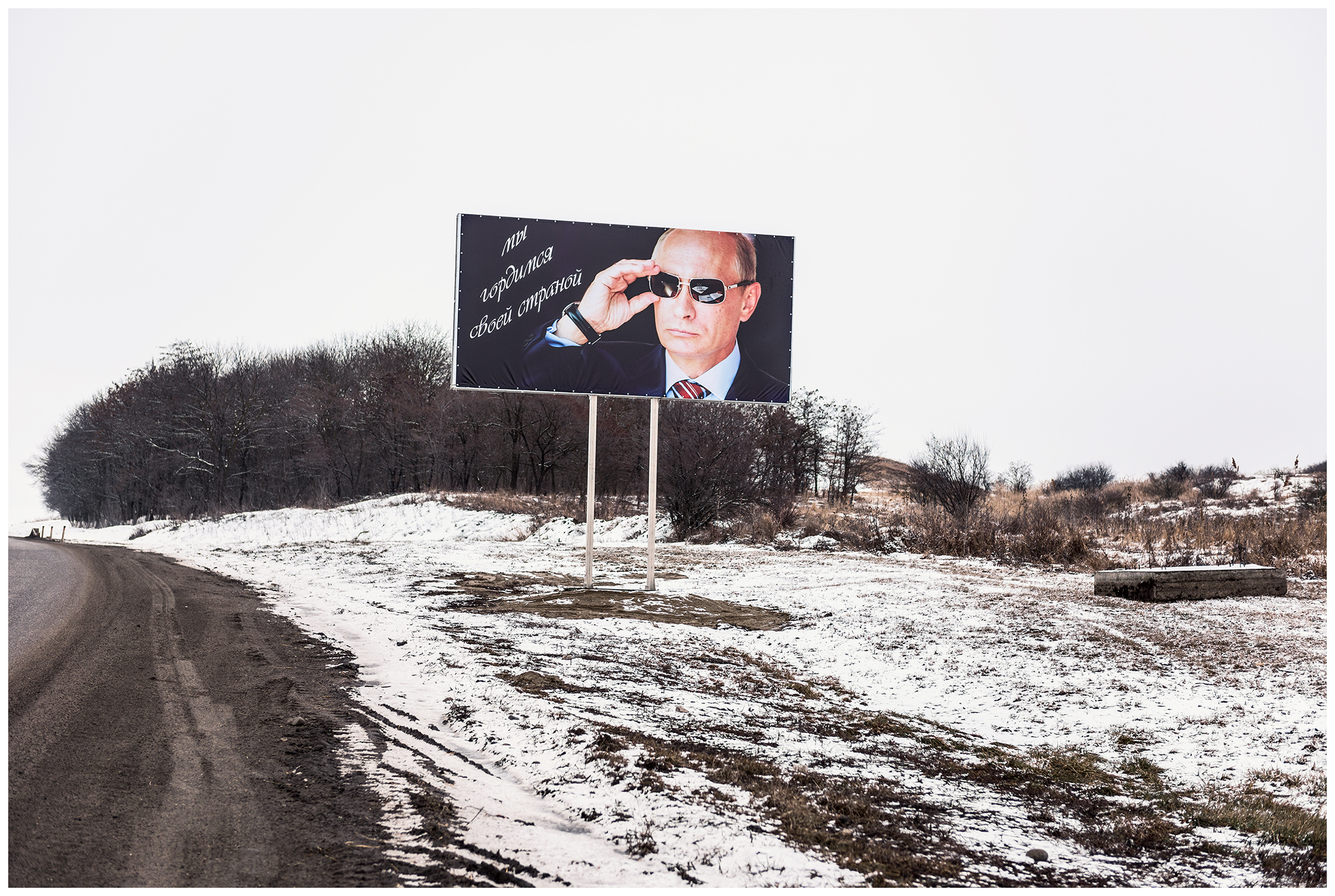 Kabardino-Balkaria, Russia. A poster with Putin and the stroke "We are proud of our country" stands at the roadside. Two hands lying on a table are reflected in the glasses. The Russian president grabs the glasses with a third hand.
