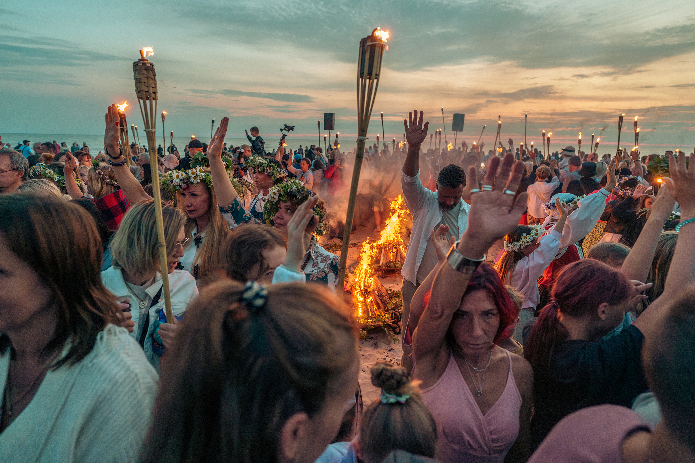 A large crowd celebrates the traditional Latvian Midsummer festival (Jāņi) on a sandy beach in Latvia at dusk, wearing elaborate flower and oak leaf crowns, holding burning torches, and raising their hands around a huge, blazing bonfire near the Baltic Sea.