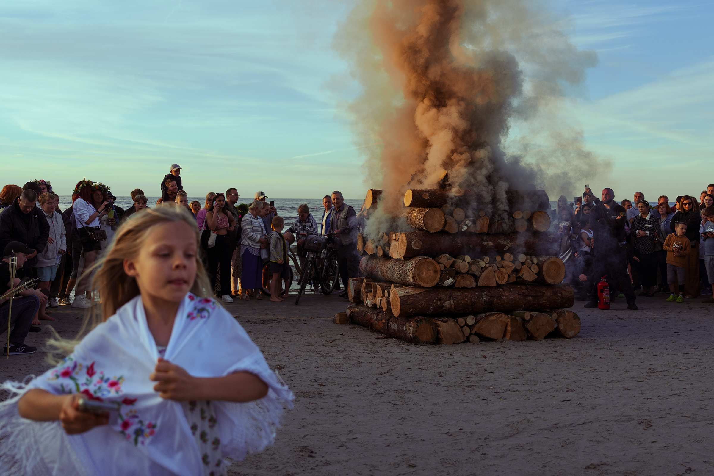 A large crowd is gathered on a sandy Baltic beach in Latvia to watch the traditional Jāņi bonfire (Jāņu ugunskurs), which is a massive stack of logs beginning to smoke, while a blurred young girl in a traditional Latvian folk costume blouse is in the foreground.