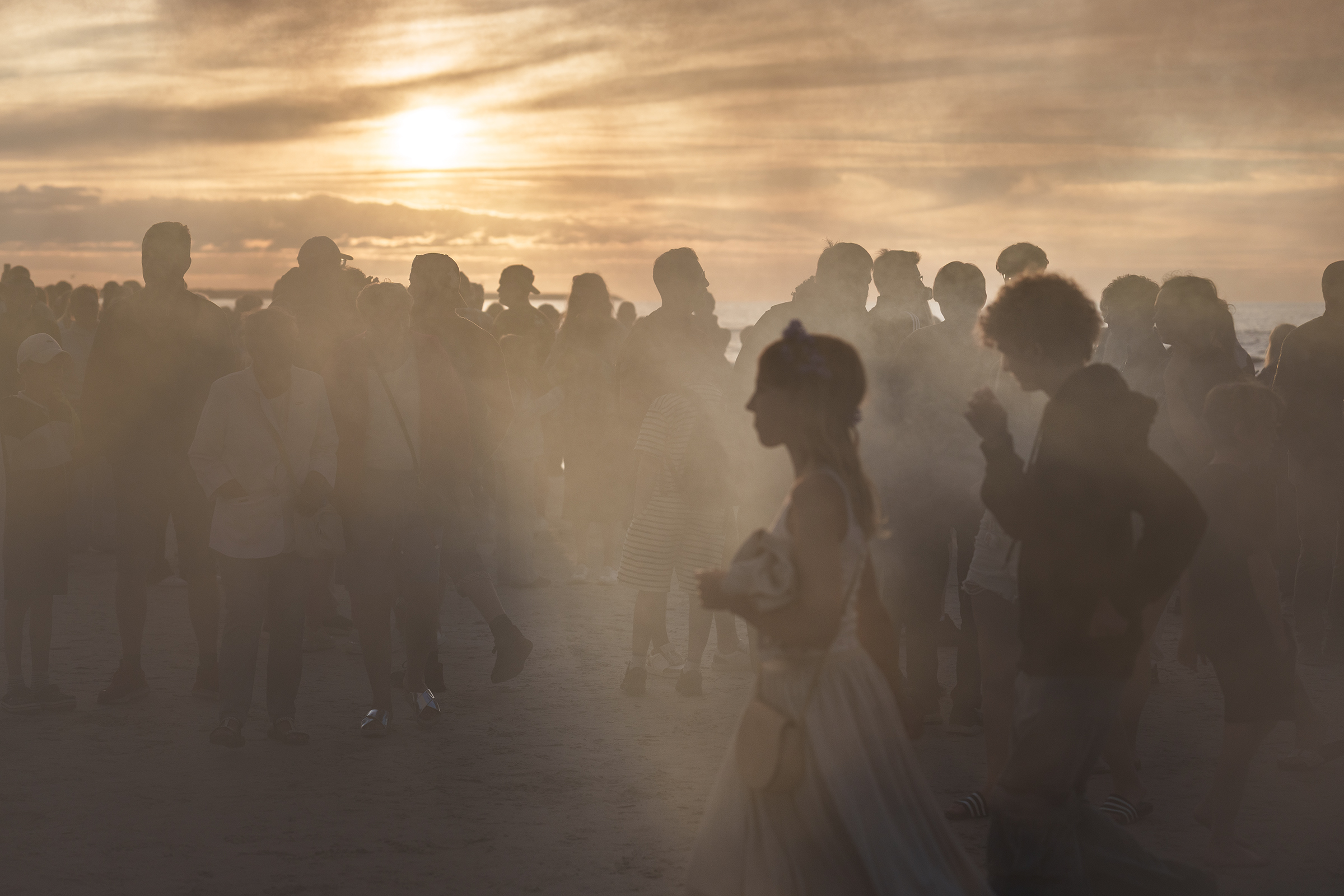 A crowd of people silhouettes on a Latvian beach gather near the Baltic Sea at sunset, with light fog or haze in the air, possibly during a traditional Midsummer celebration.