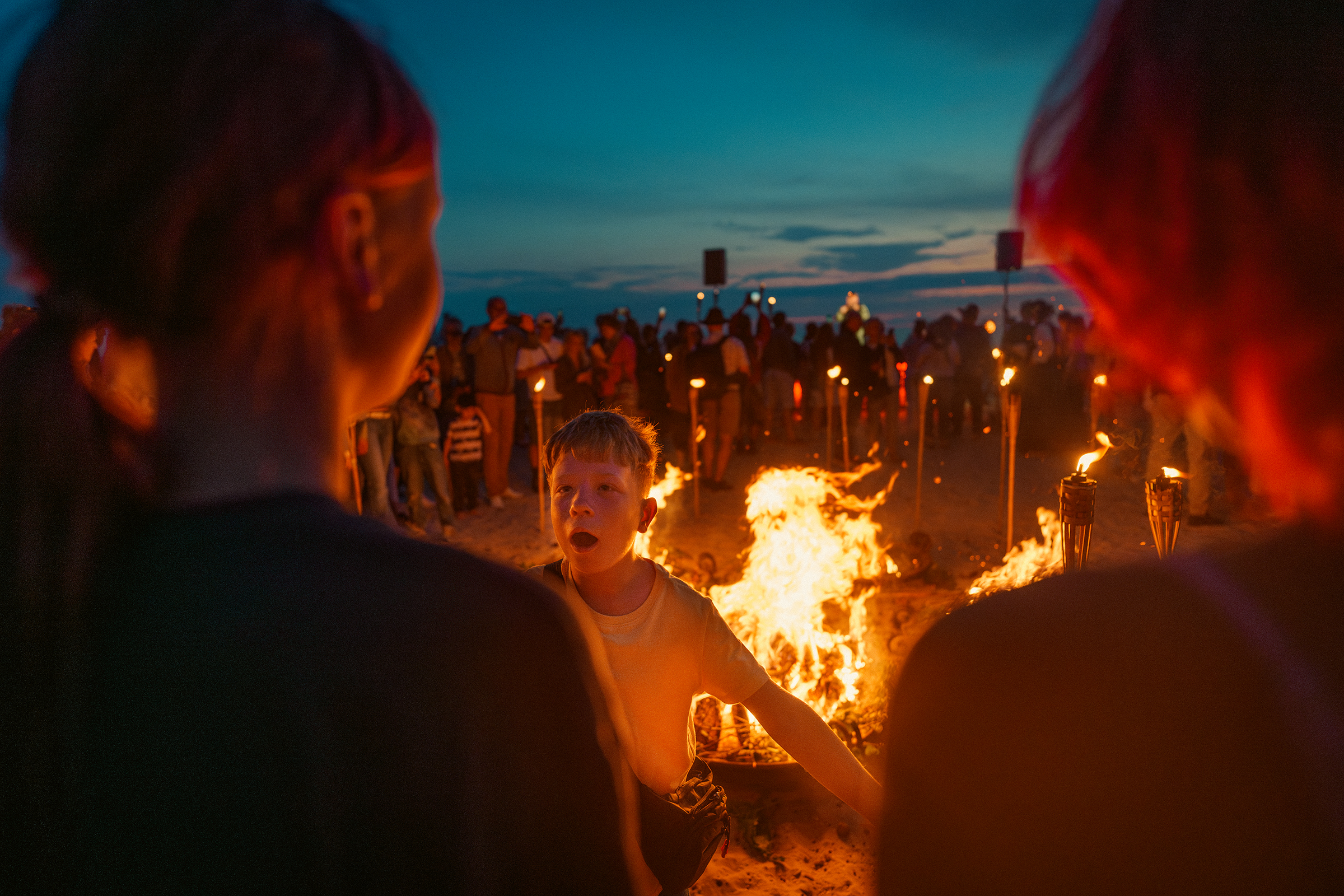 A young boy reacts in awe near a beach bonfire and torches during an evening gathering along the coast of Latvia.