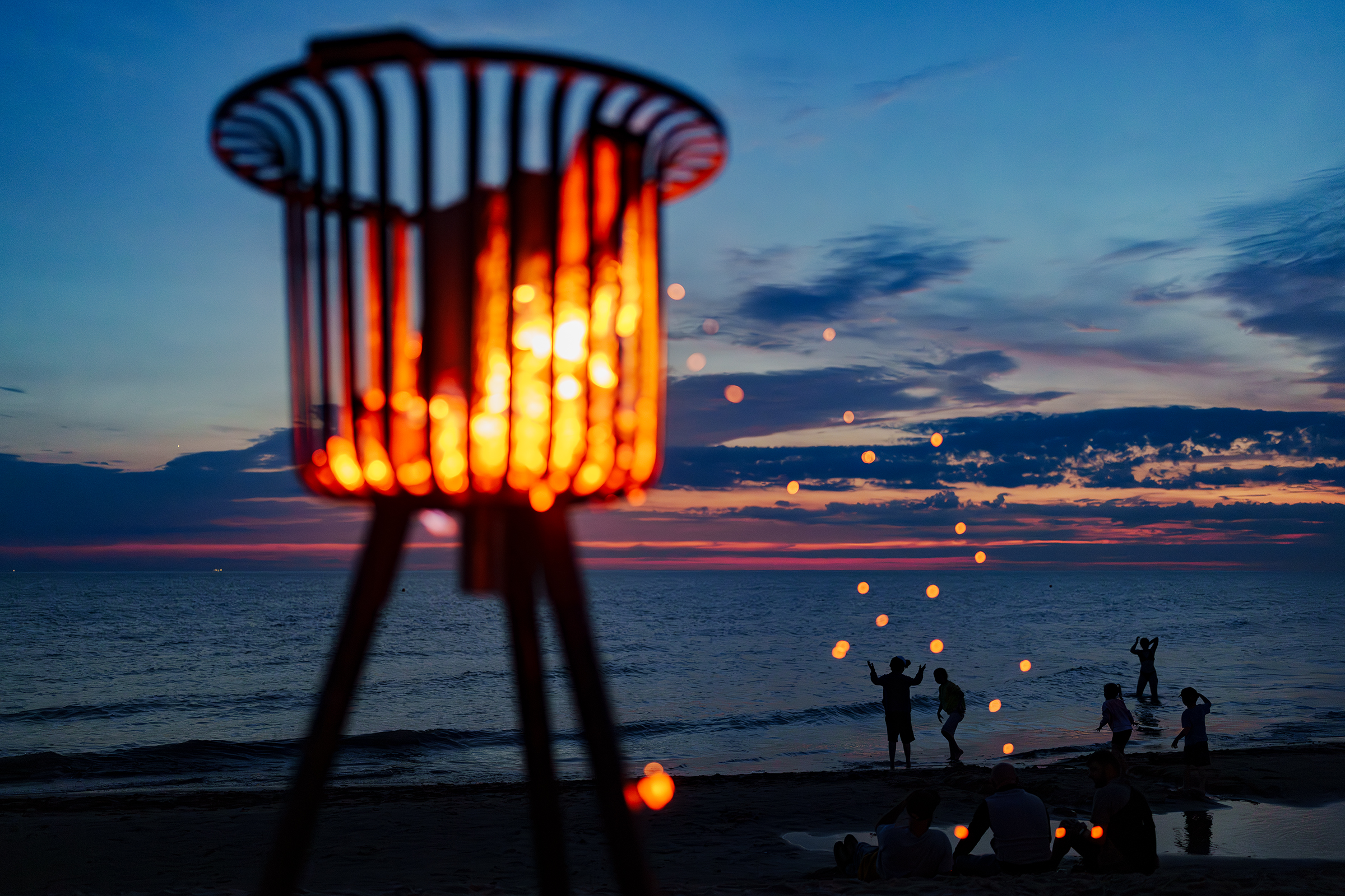 A glowing fire basket stands in the foreground as people gather along a beach in Latvia at sunset.