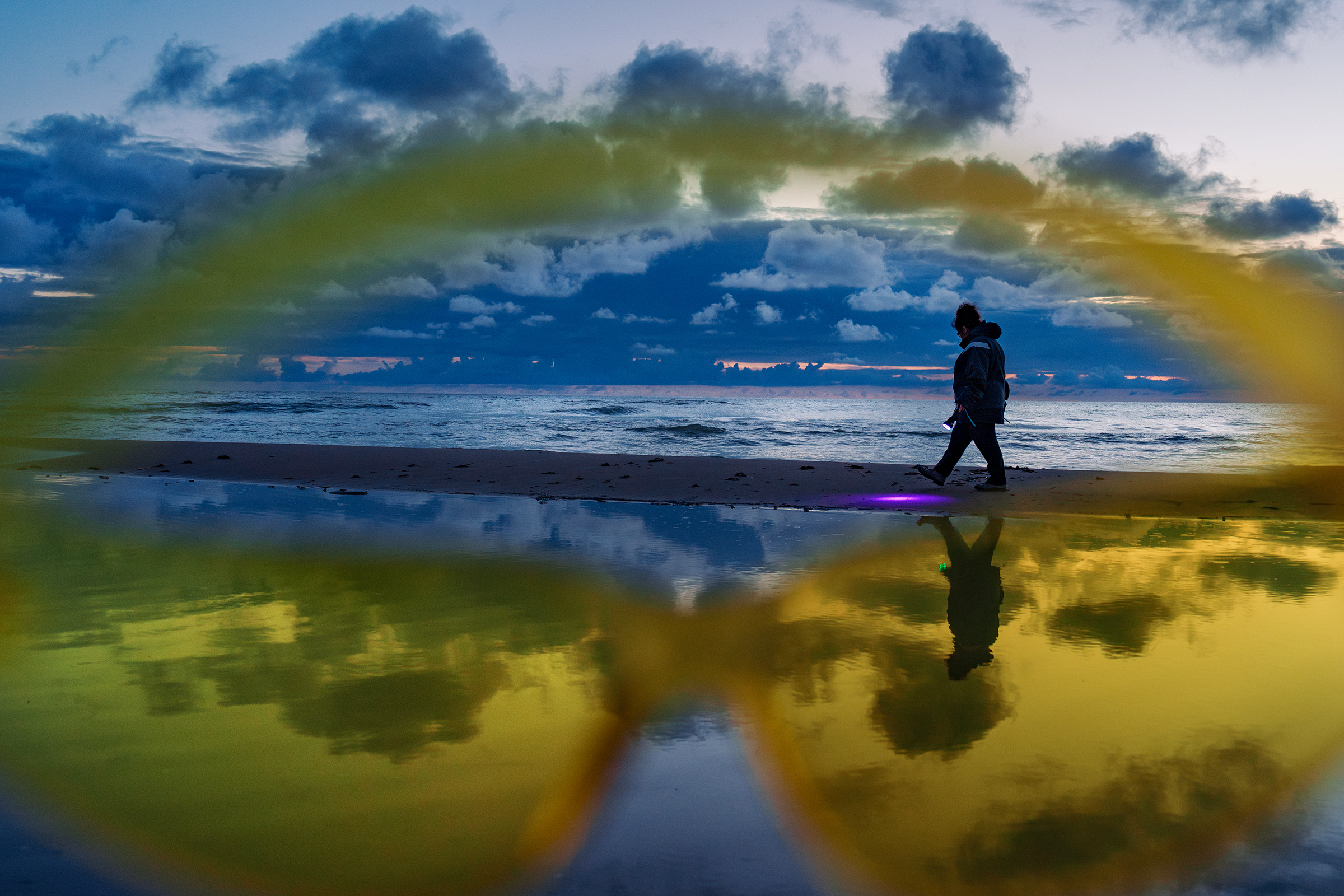 A person holding a UV flashlight walks along a beach in Latvia at dusk, framed by yellow tinted glasses in the foreground.
