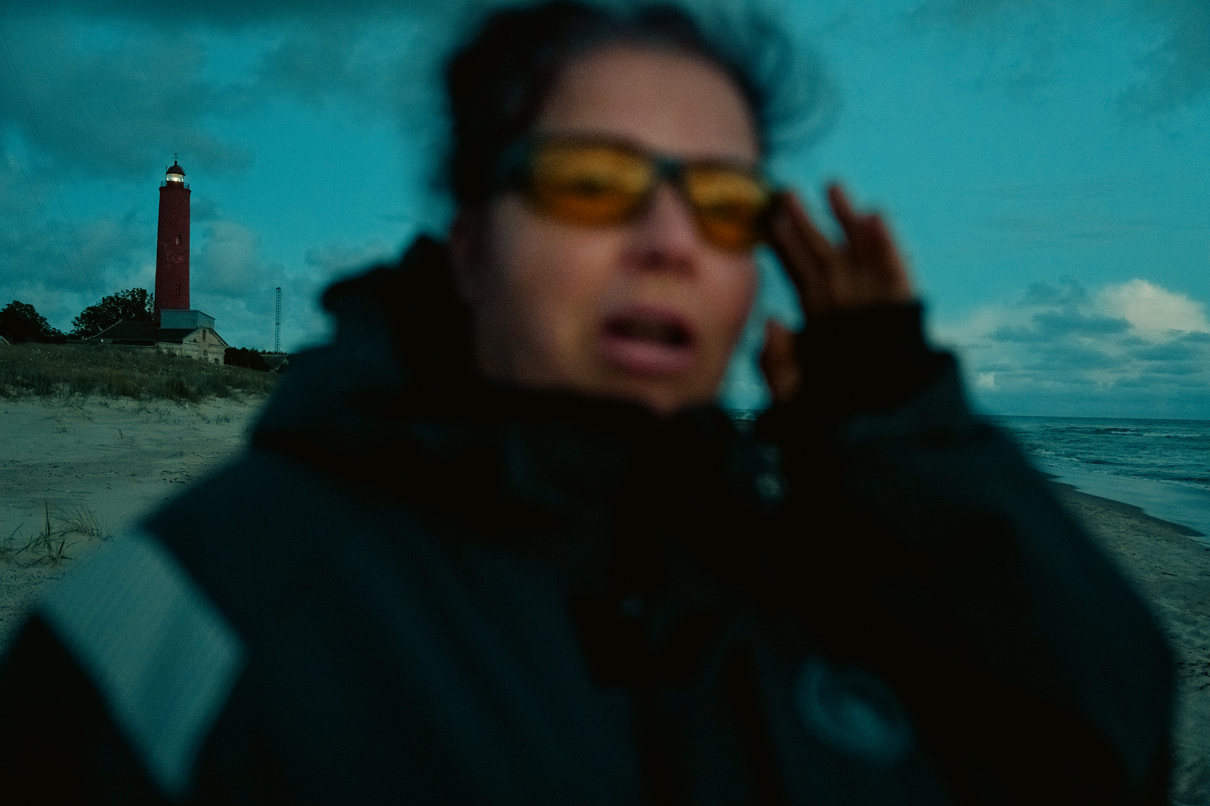 A close-up of a blurred person wearing yellow glasses on a beach in Latvia with a red lighthouse lit up in the background.