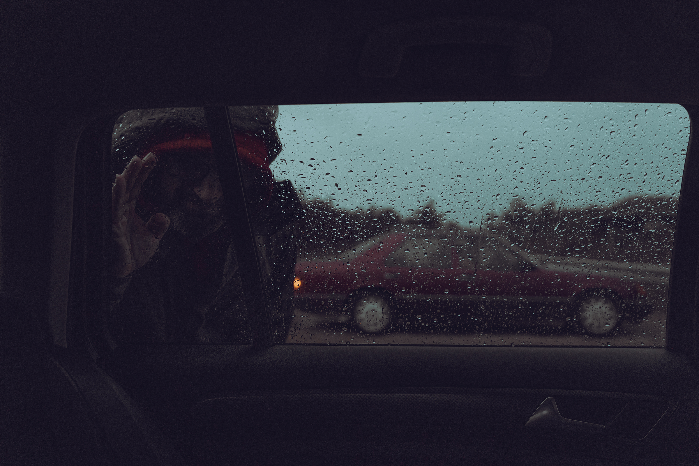 A view from inside a car on a rainy day in Latvia showing a person outside pressing their hand against a rain-covered window.