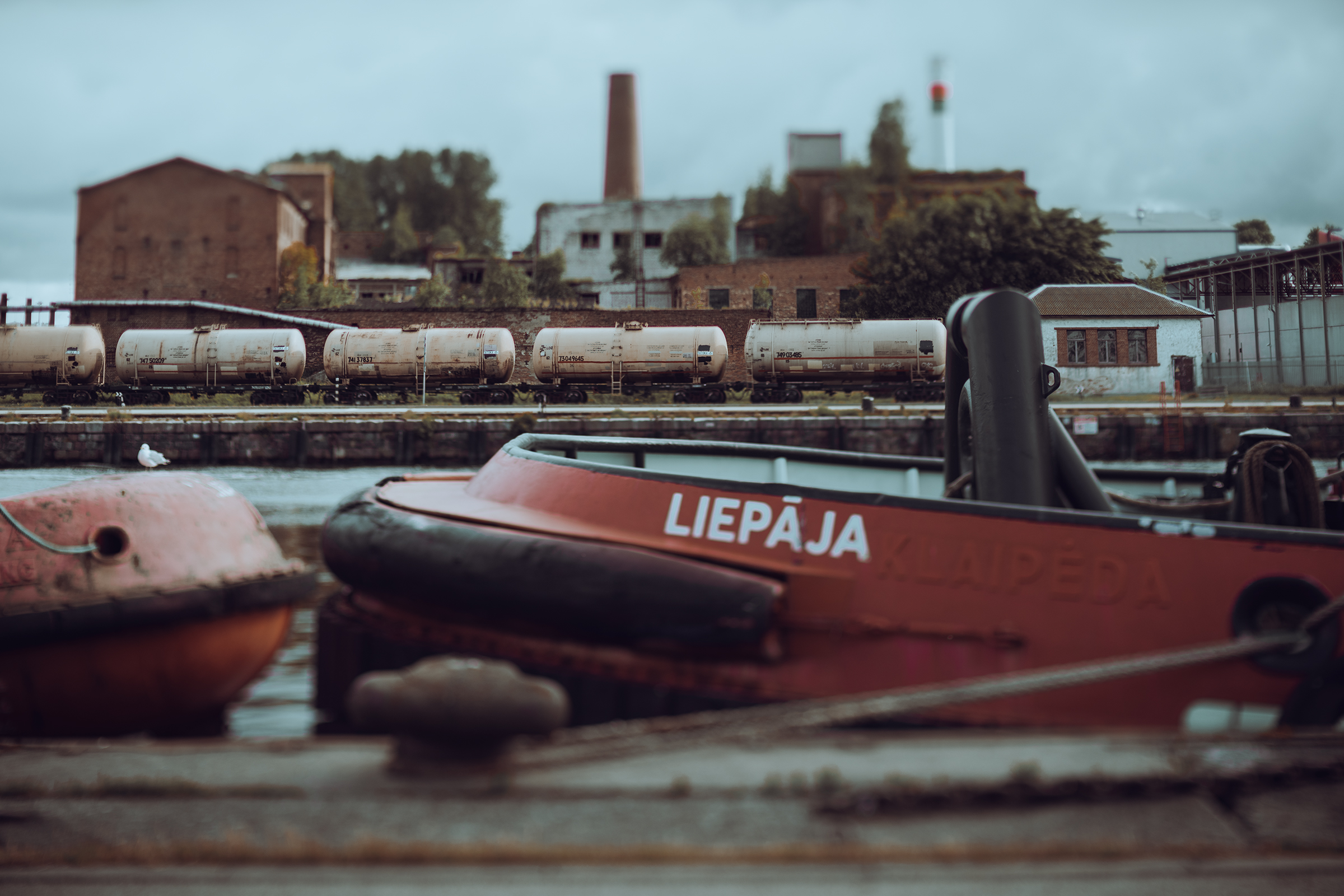 A boat displaying the name of the city Liepāja docked at an industrial port in Latvia with train tanker cars in the background.