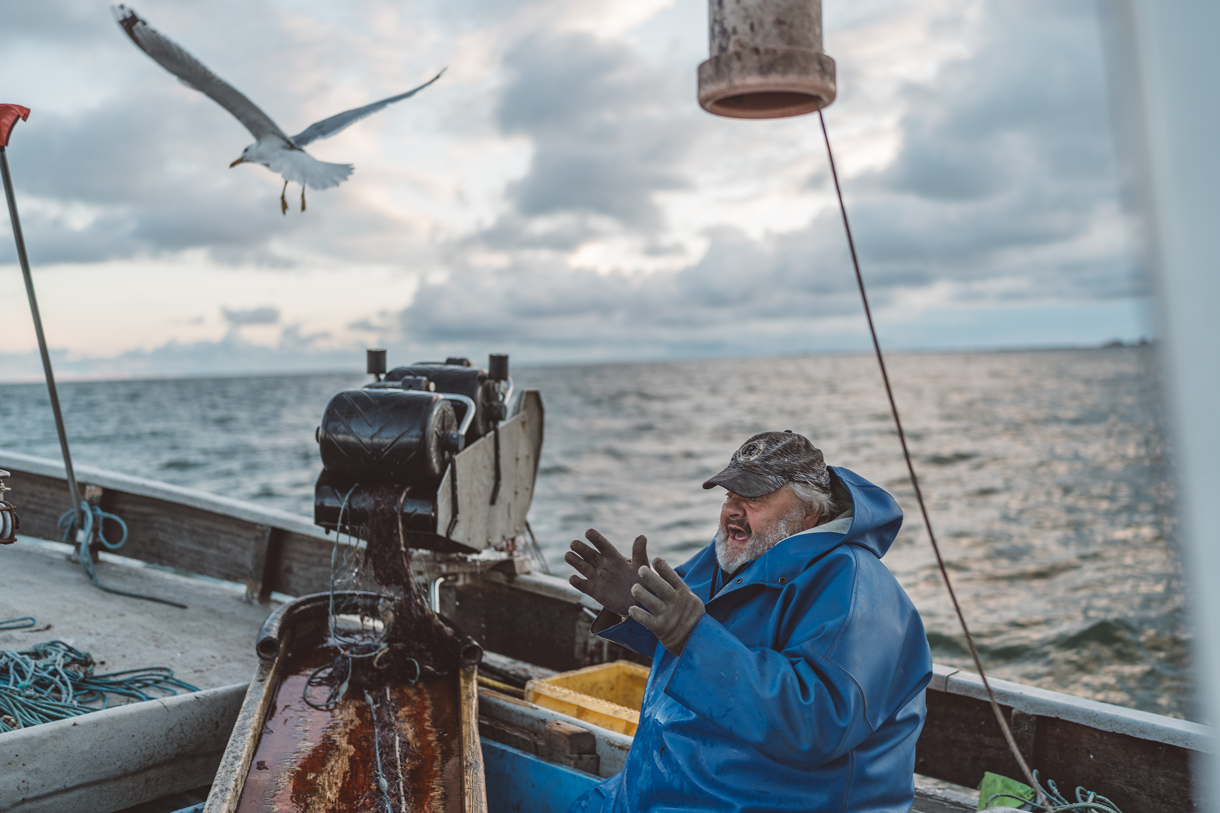 A fisherman in a blue raincoat gestures on a boat off the coast of Latvia as a seagull flies overhead.