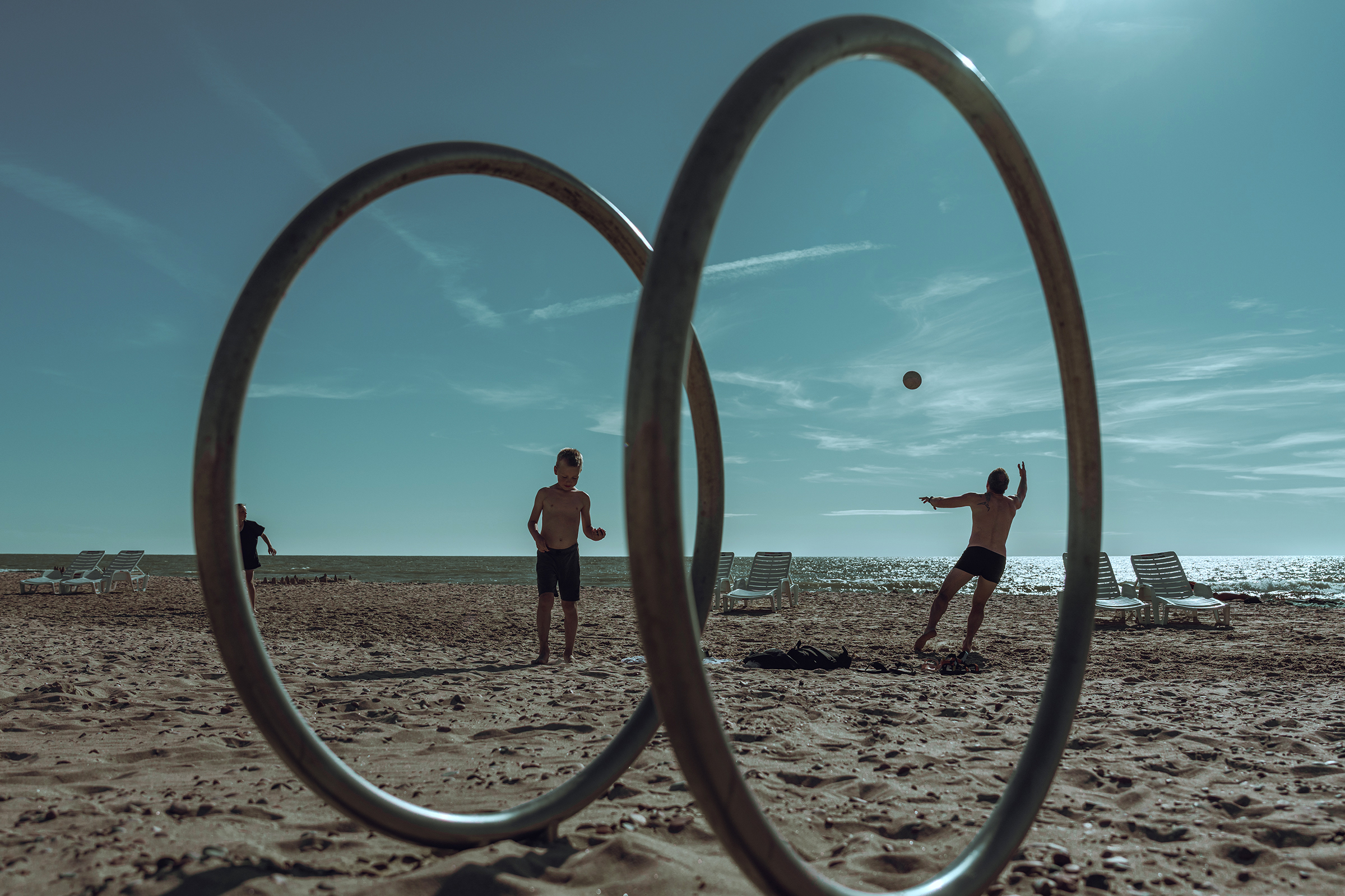 People play ball on a sunny beach in Latvia, framed by large metal gymnastic rings installed in the sand.