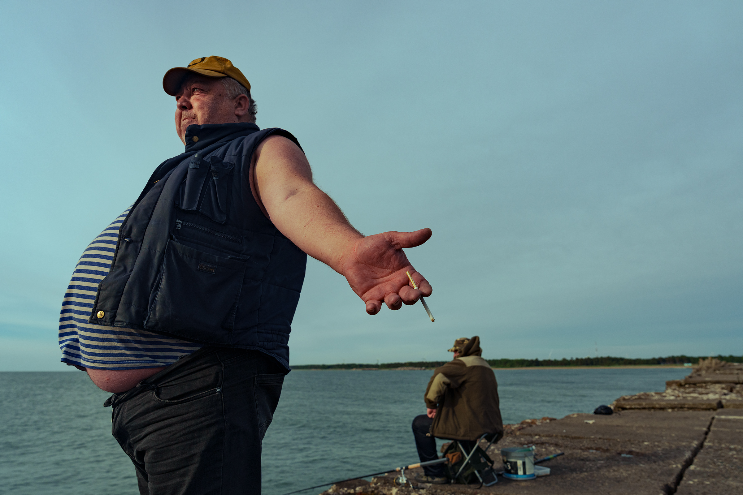 A man wearing a yellow cap and striped shirt holds out a cigarette while standing on a concrete pier in Latvia, with another angler sitting in the background.