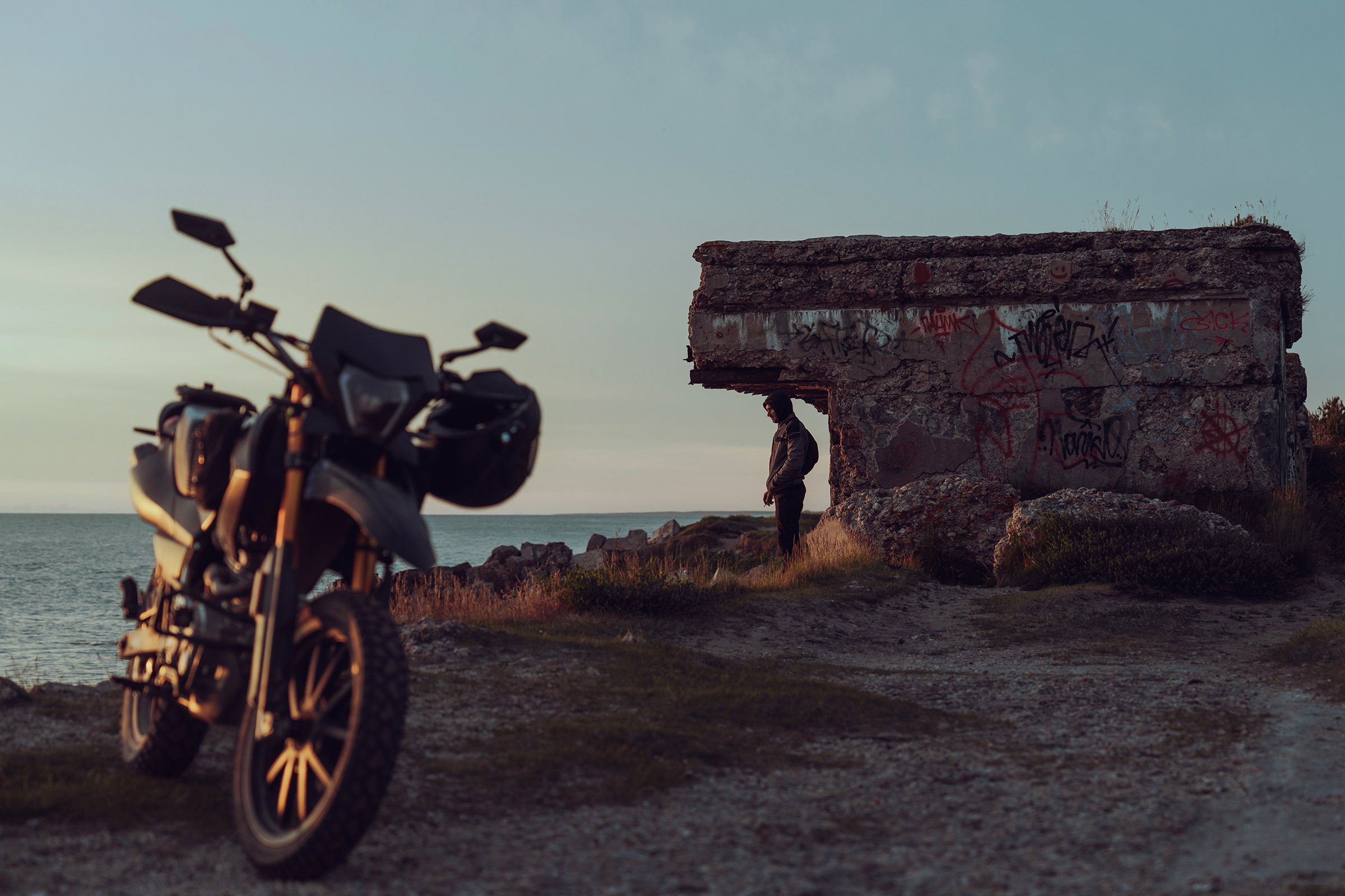 A motorcyclist stands under the overhang of a graffiti-covered concrete bunker by the sea in Latvia, with a motorcycle parked in the foreground.