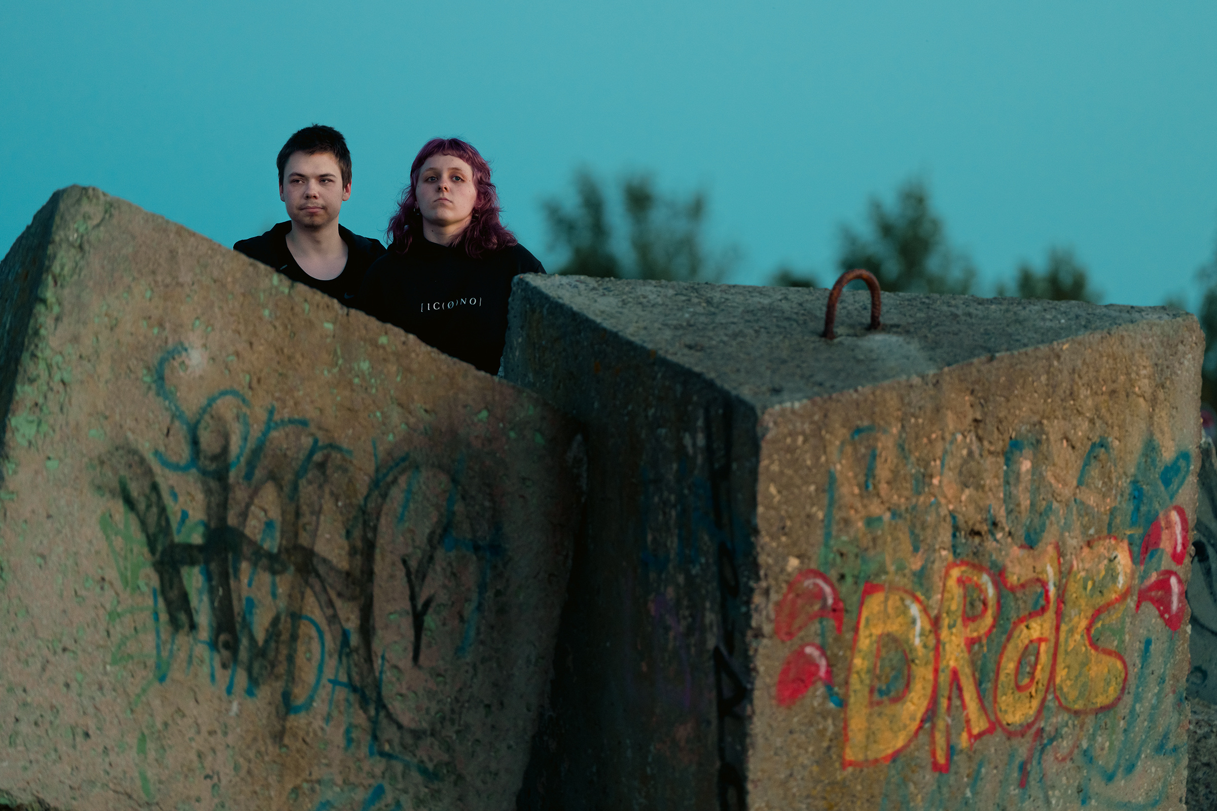 Two young people stand behind large, graffiti-tagged concrete blocks along the coast in Latvia.