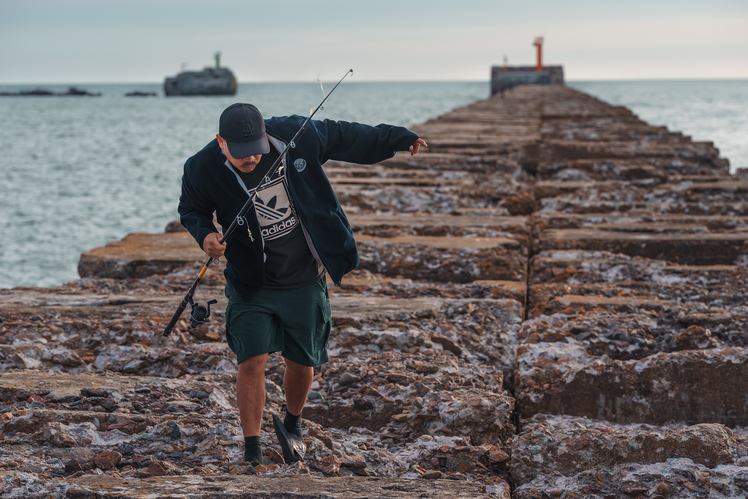 A fisherman holding a fishing rod walks carefully along a long, weathered stone pier in Latvia, with a red beacon visible at the end.