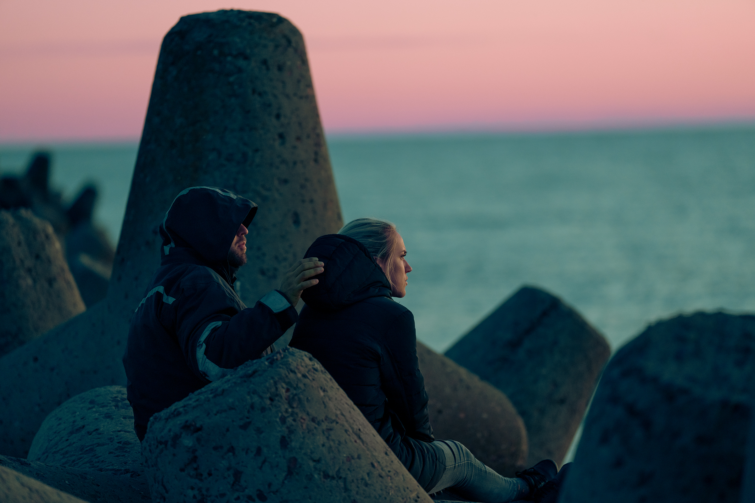 A hooded couple sits among large concrete tetrapods along the Baltic coast in Latvia during a pink and blue twilight sky.