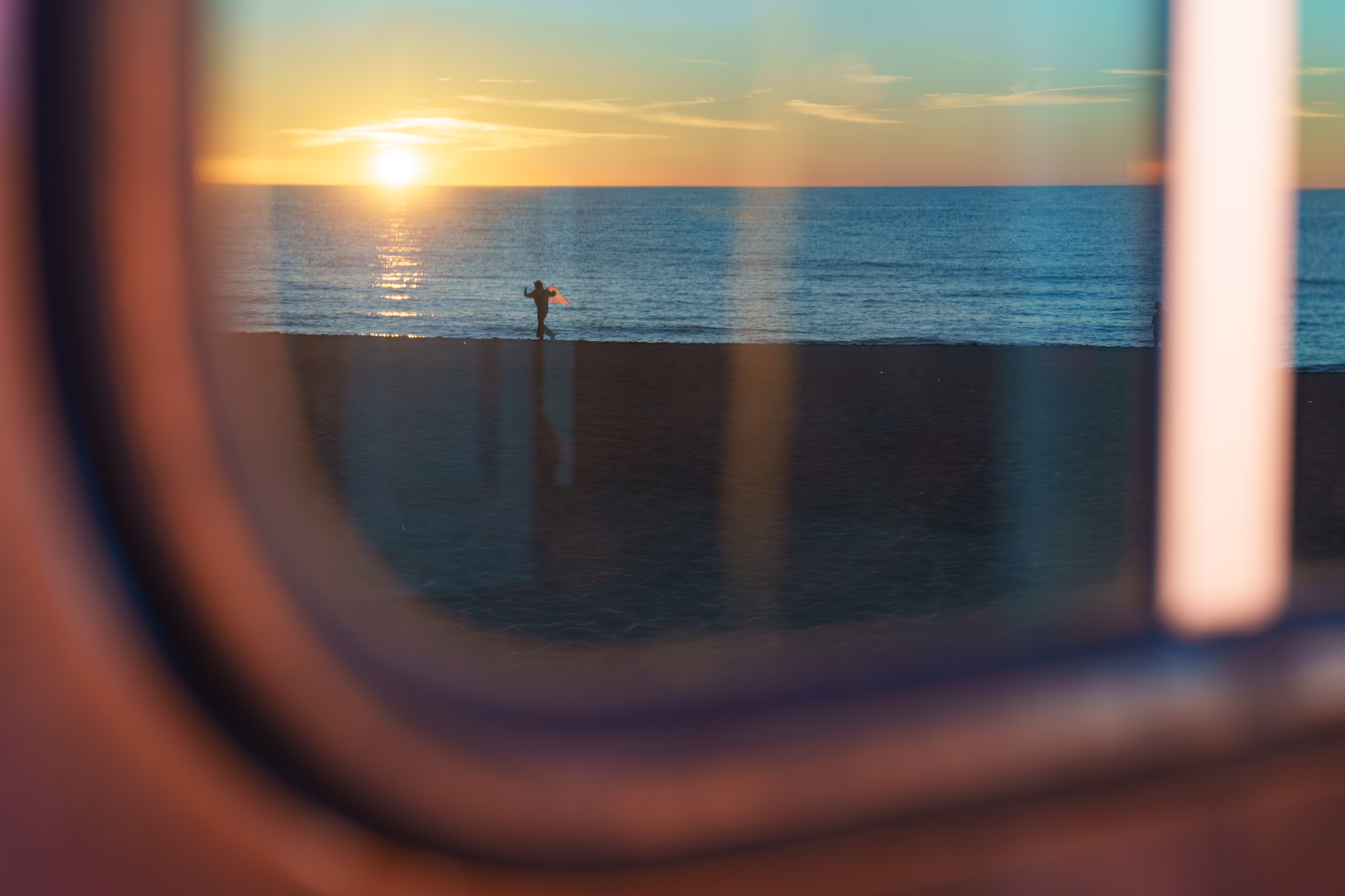 A person holding a kite walks along a sandy beach at sunset in Latvia, framed through a window.