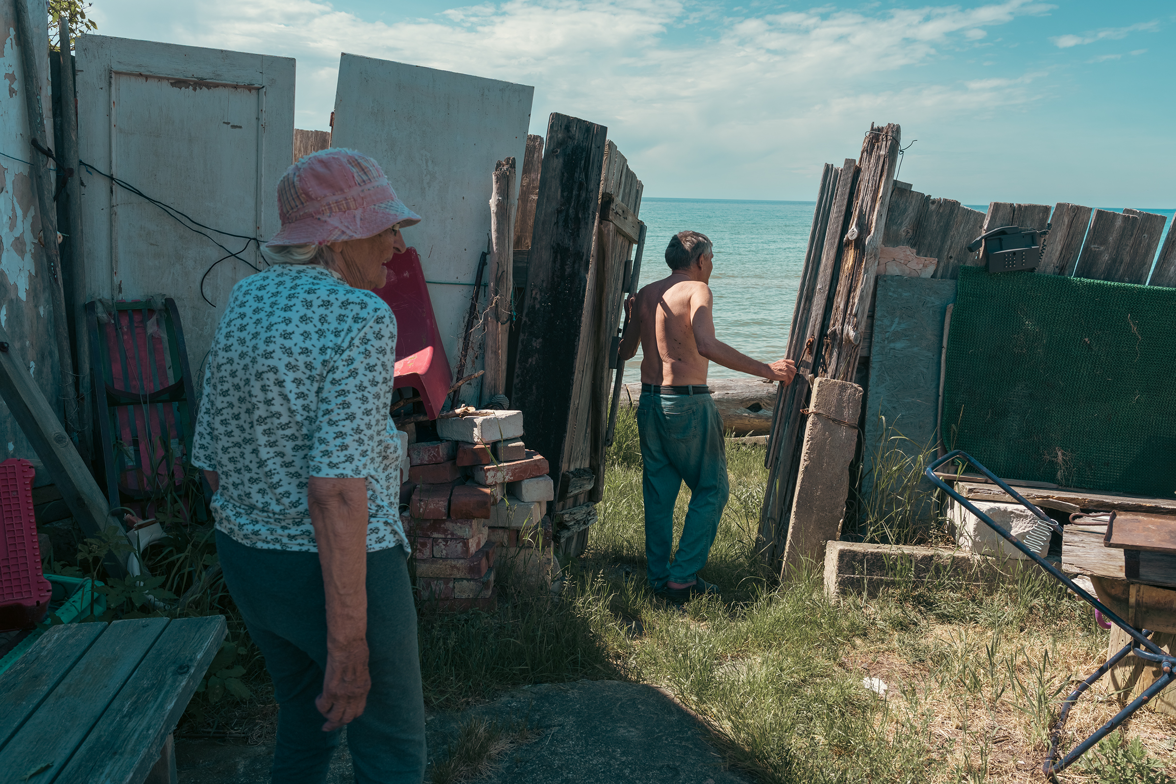 An elderly couple stands by a makeshift wooden fence near the sea in Latvia.