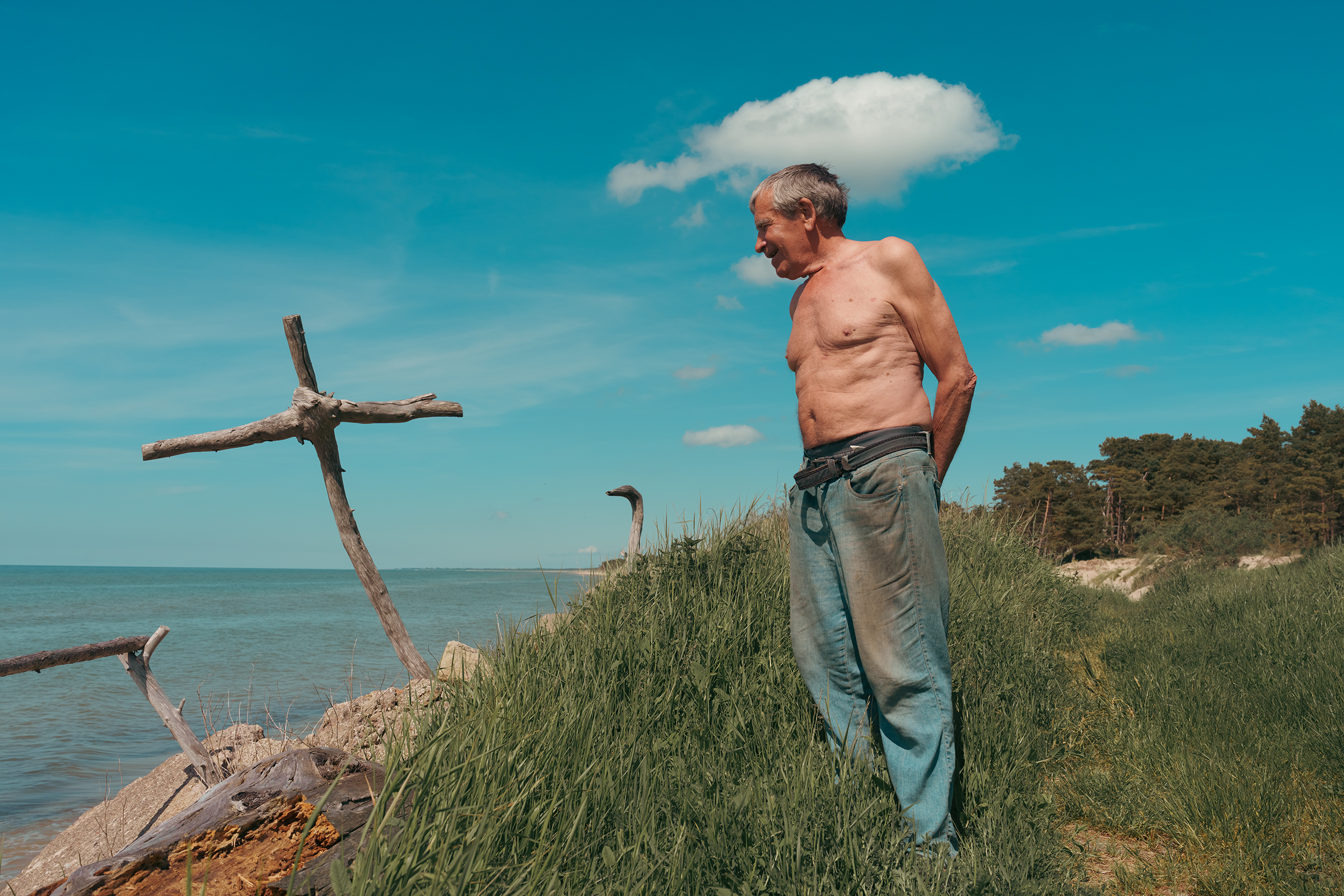 A shirtless man stands on a grassy coastal bluff in Latvia looking towards a wooden cross made of driftwood by the sea.