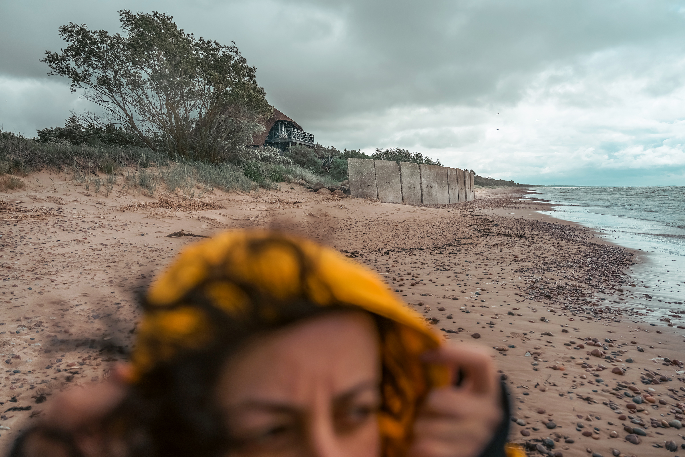 A woman wearing a yellow hooded jacket appears blurred in the foreground of a windy beach in Latvia, with concrete sea barriers and a building visible along the dunes under an overcast sky.