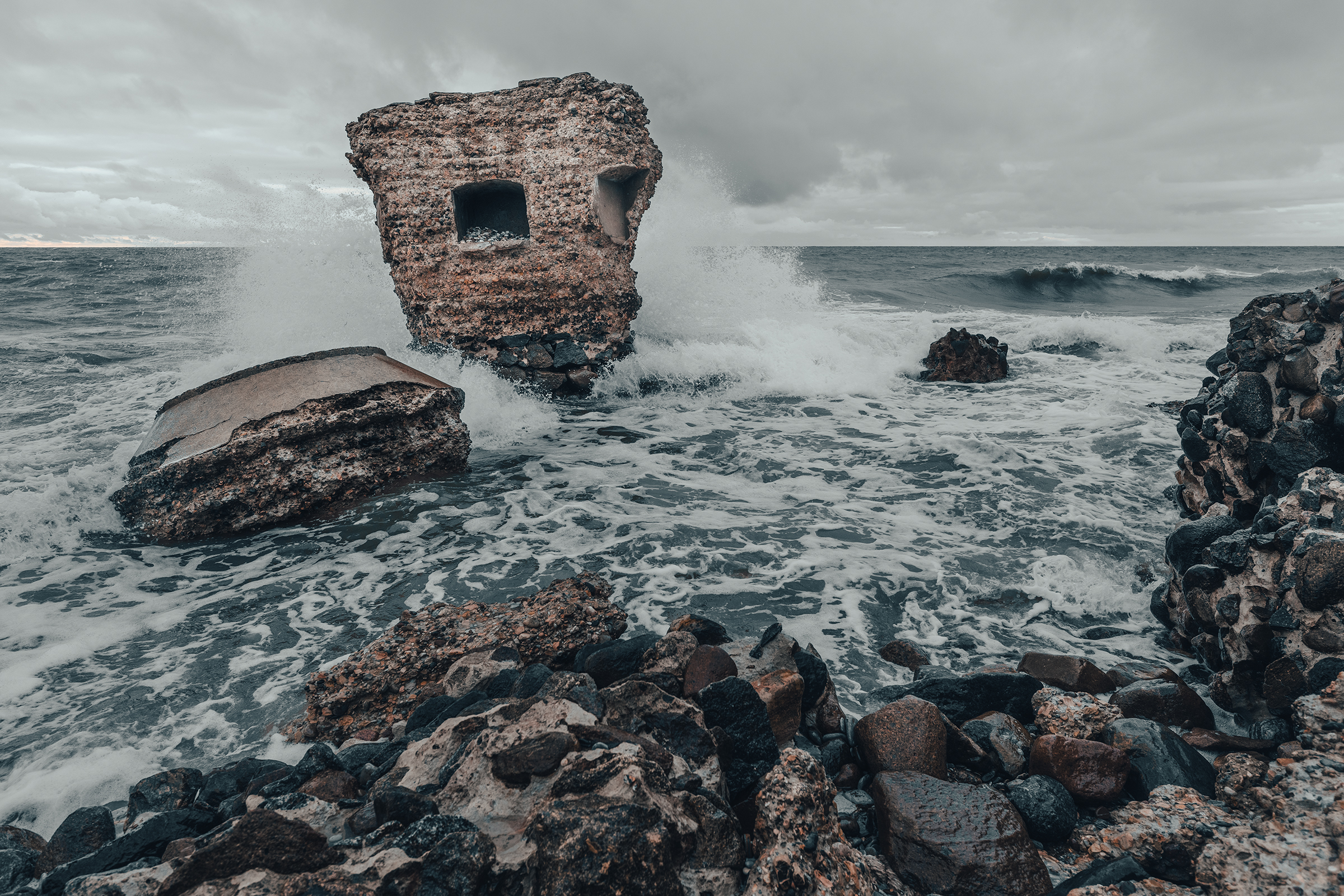 Waves crash against a partially submerged, crumbling concrete bunker offshore along the Baltic coast in Latvia under a stormy grey sky.