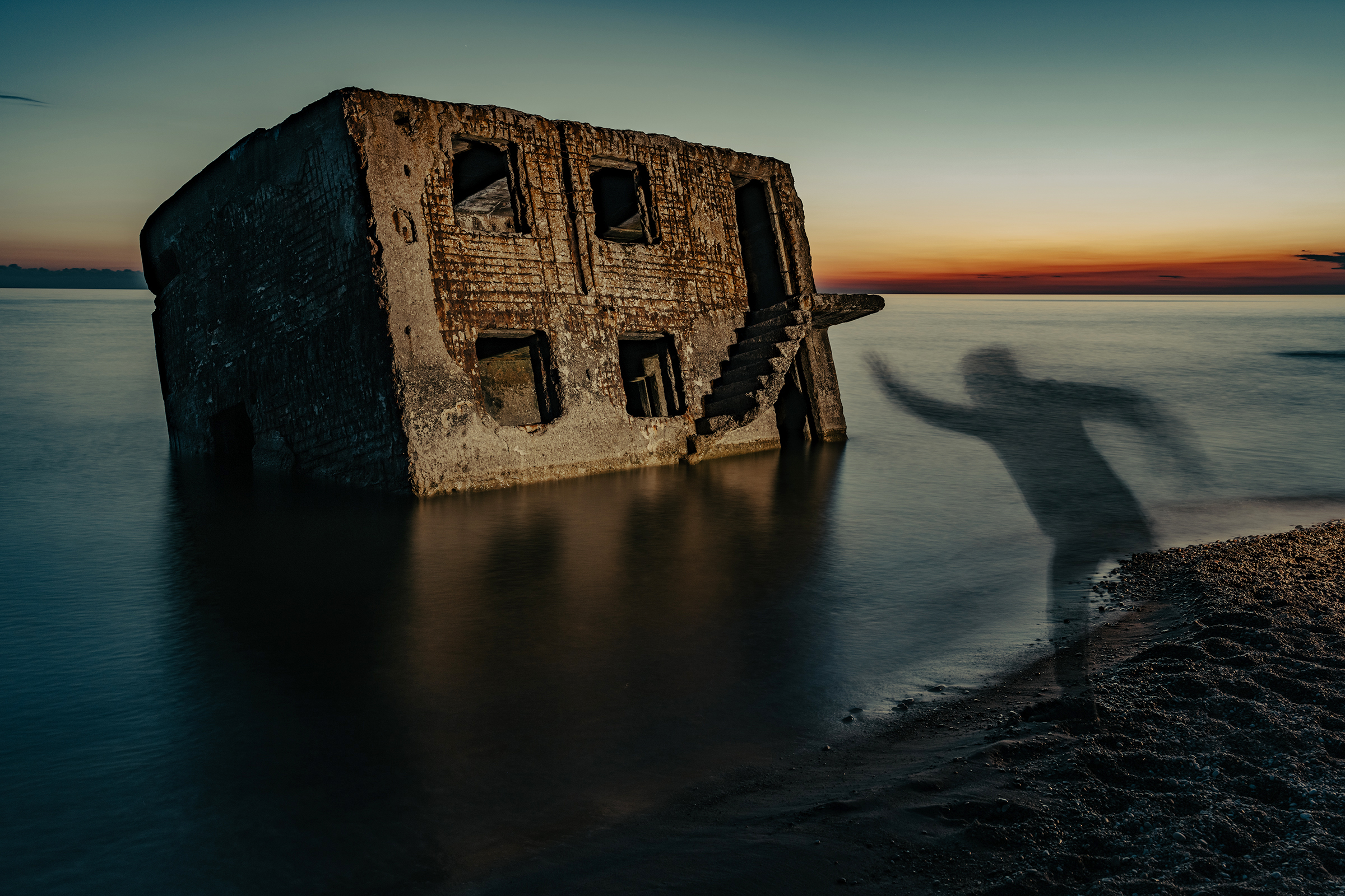 A long exposure photo captures a motion-blurred figure running along the shore next to a tilted, abandoned concrete bunker resting in the sea under a sunset sky in Latvia.