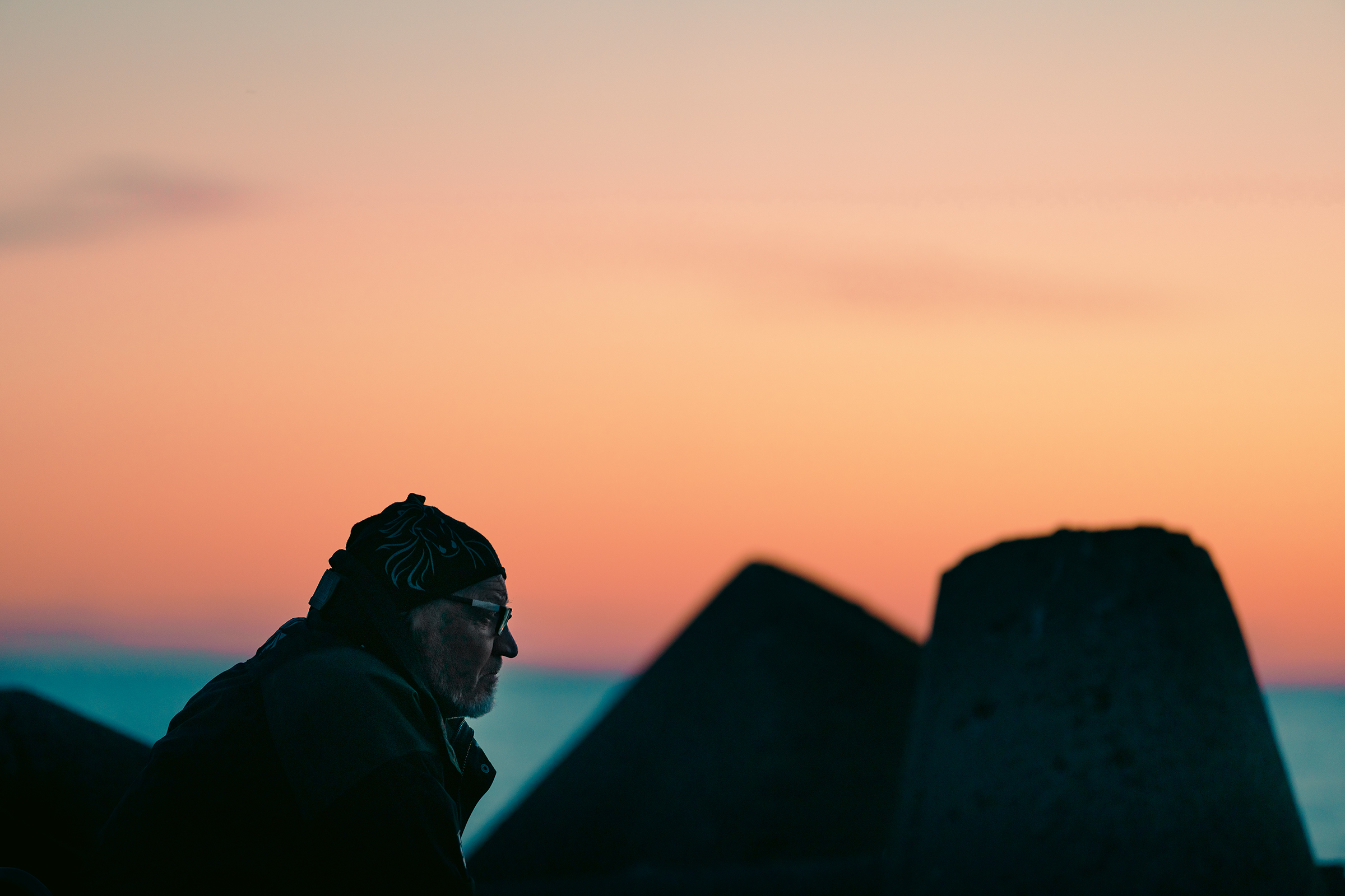 A side profile of an older man wearing a beanie and glasses, silhouetted against a vibrant orange and pink sunset behind concrete breakwater blocks in Latvia.