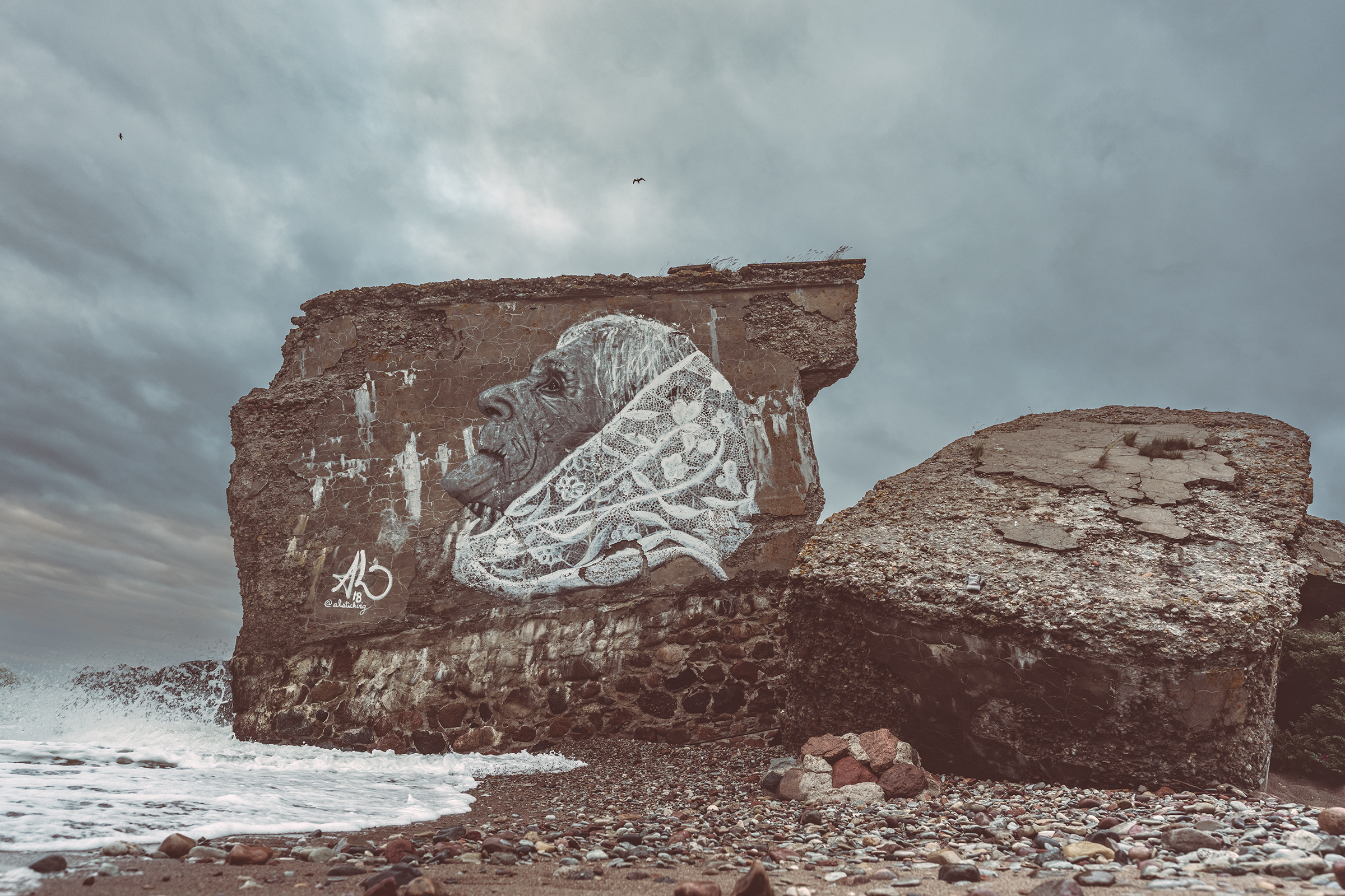 A large painted mural of an elderly woman wearing a headscarf adorns the wall of a crumbling concrete bunker structure on a stony shore in Latvia under a cloudy sky.
