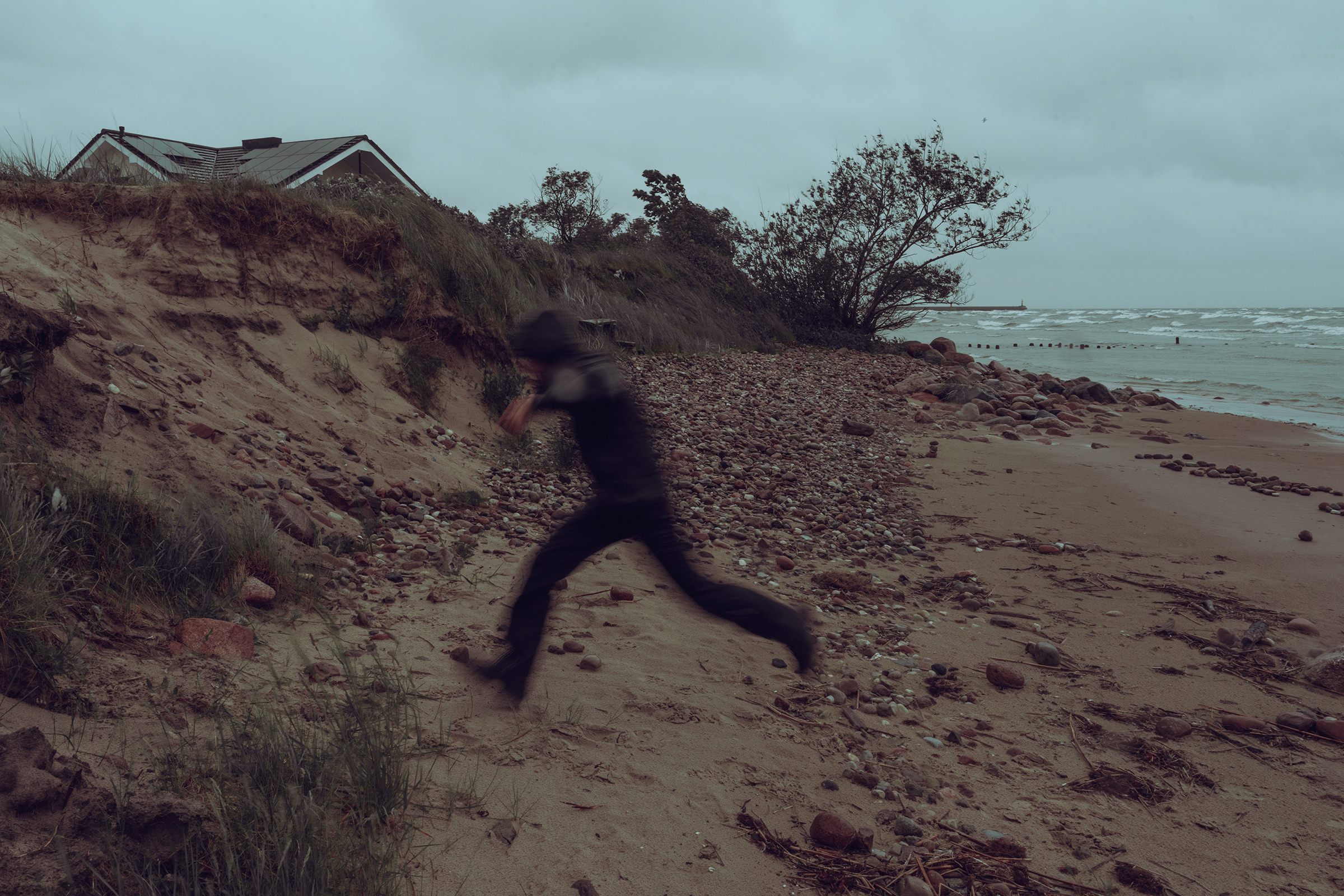 A motion-blurred figure runs along an eroded sandy slope next to a stony beach in Latvia, with a house visible at top left under an overcast sky.
