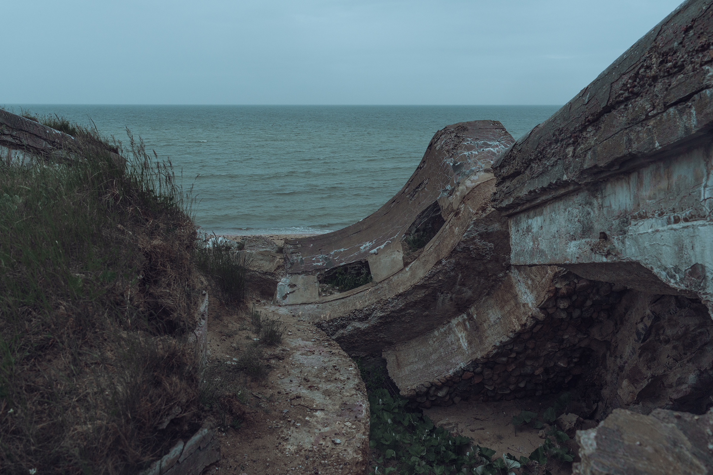 Cracked and collapsed concrete military bunker ruins sit on an eroded coastal cliff overlooking the calm Baltic Sea in Latvia.