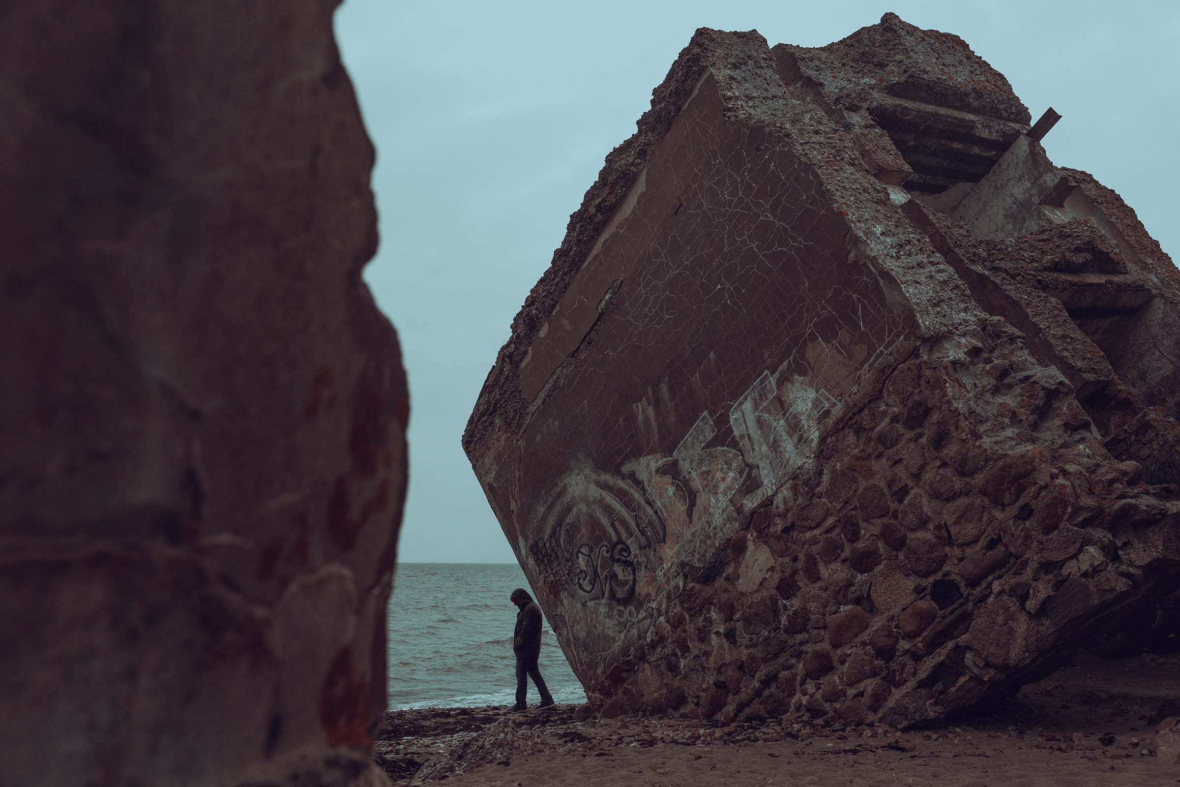 A hooded person walks along the beach next to a massive, tilted concrete bunker remnant on the coast of Latvia under an overcast sky.