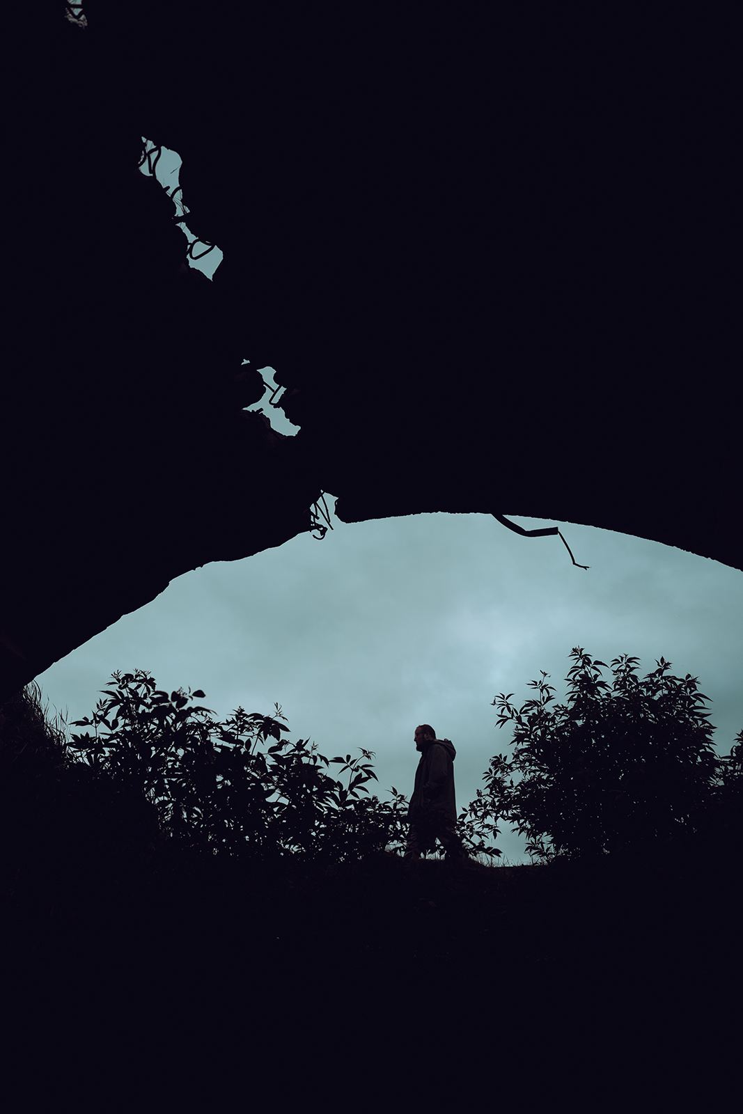 A silhouetted person walks past bushes, framed by the dark archway of a damaged concrete bunker structure in Latvia under a cloudy twilight sky.