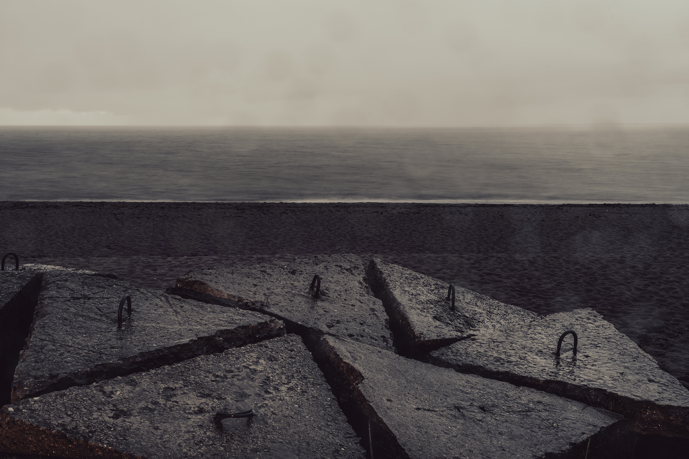 Large triangular concrete breakwater blocks with metal lifting loops sit along a dark, moody coastline facing a calm ocean horizon in Latvia.