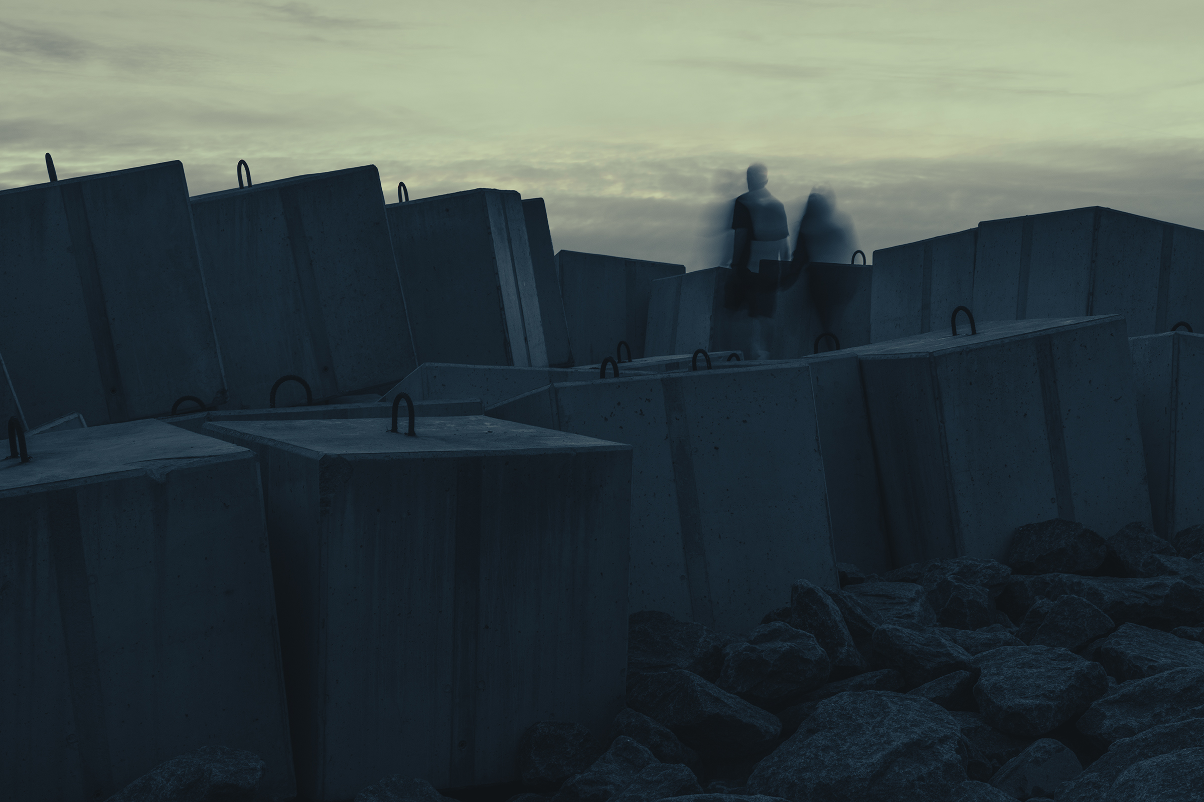Two motion-blurred figures stand among massive concrete sea wall blocks on a rocky coast in Latvia under a muted evening sky.