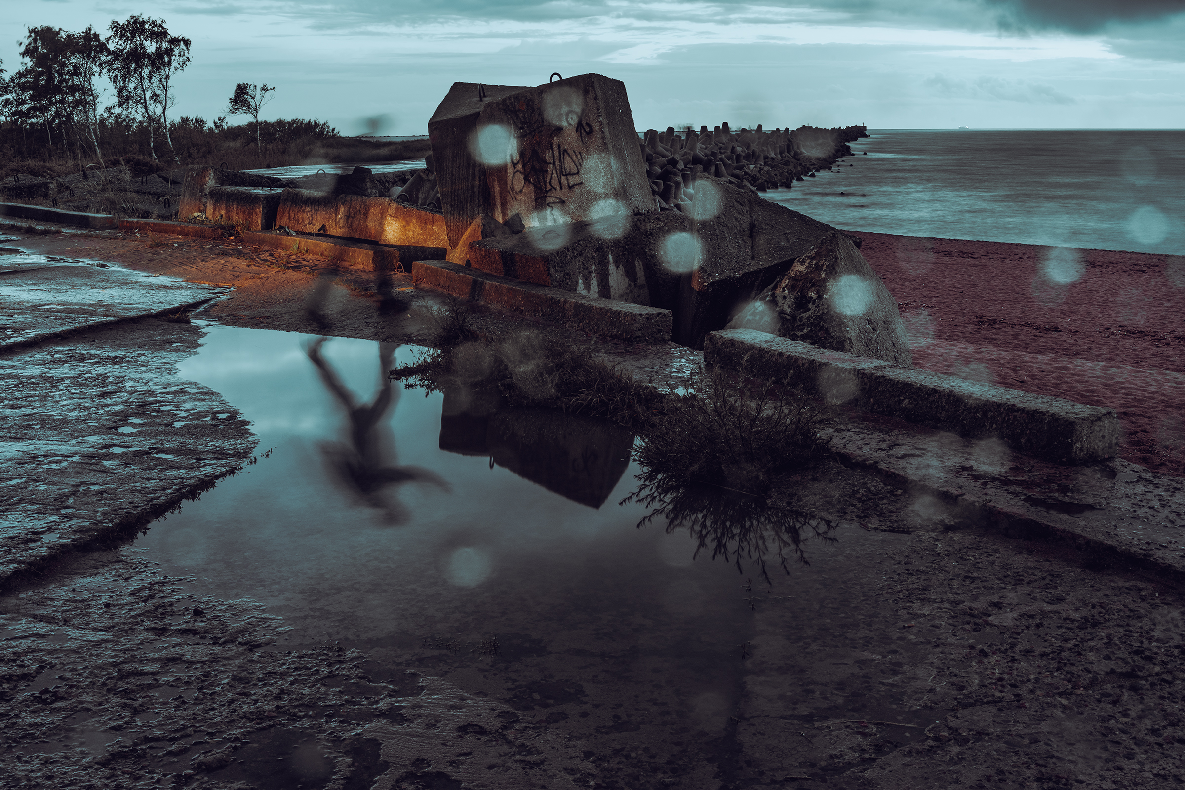 A long exposure photo captures water droplets and the reflection of a motion-blurred figure in a puddle near concrete coastal defense structures and a pier extending into the Baltic Sea in Latvia.