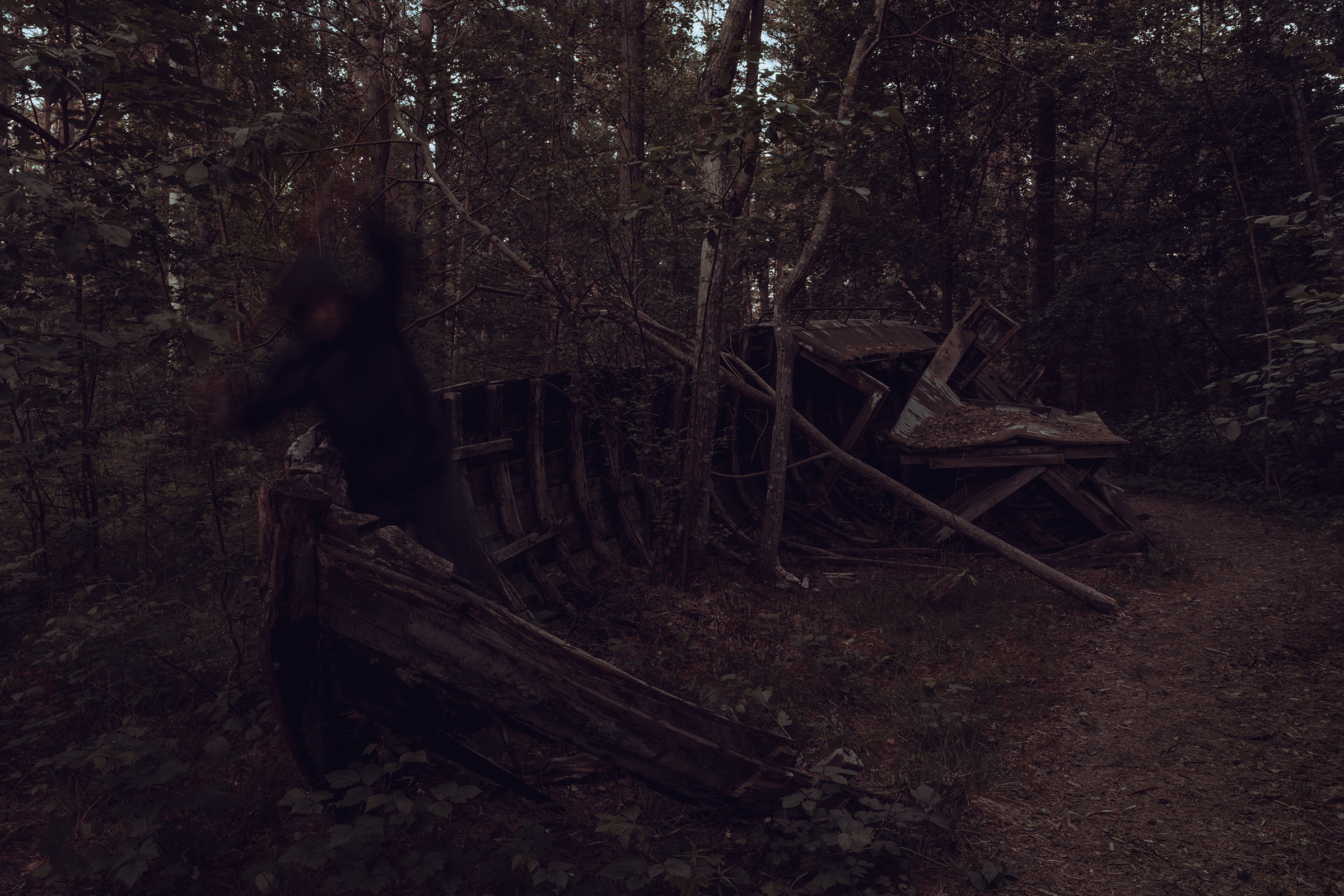 A motion-blurred hooded figure moves past the wooden ribs of a decaying ship graveyard hidden in a dense, dark forest near the coast in Latvia.