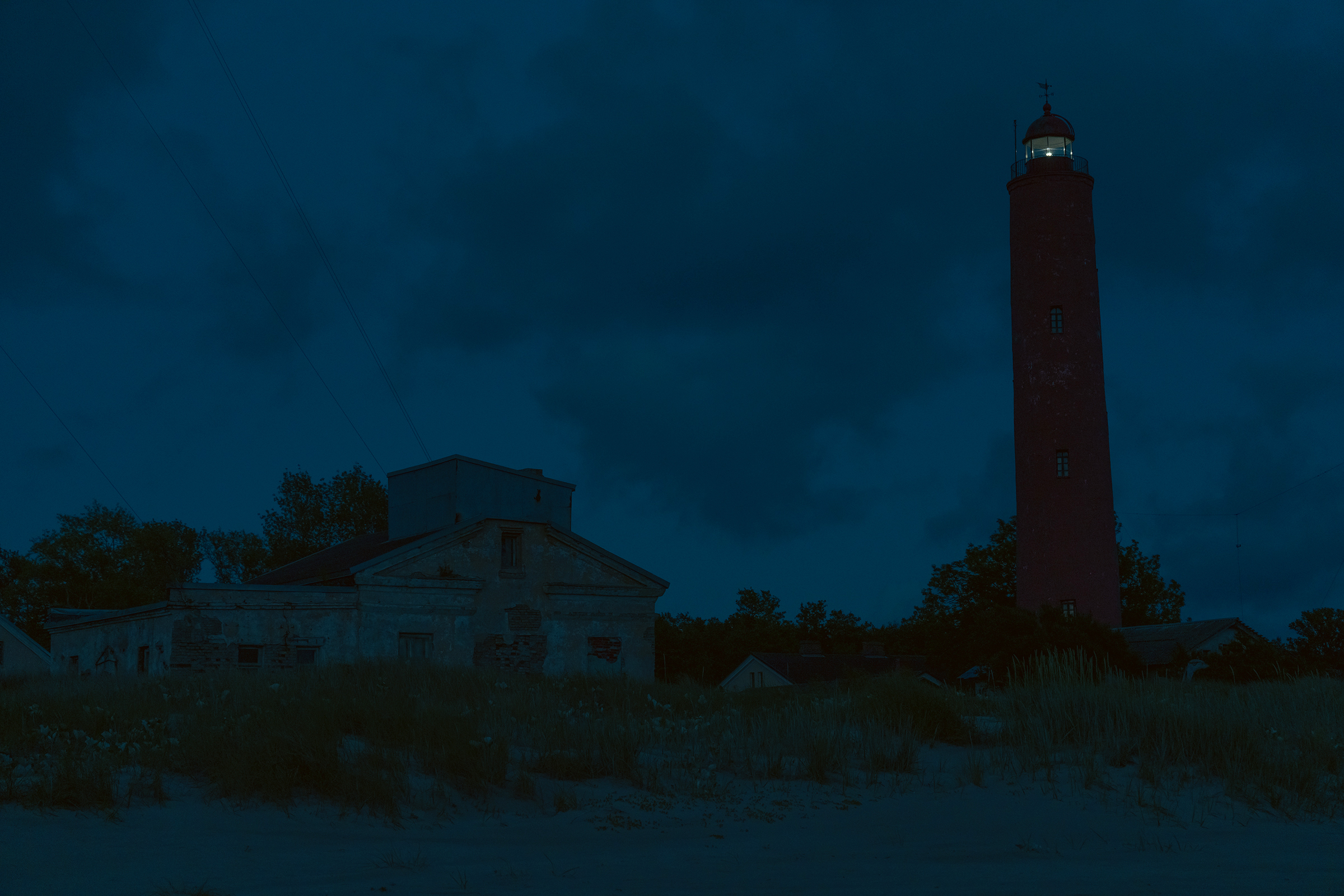 A tall red brick lighthouse with its beacon illuminated stands against a deep blue night sky next to old coastal buildings in Latvia.